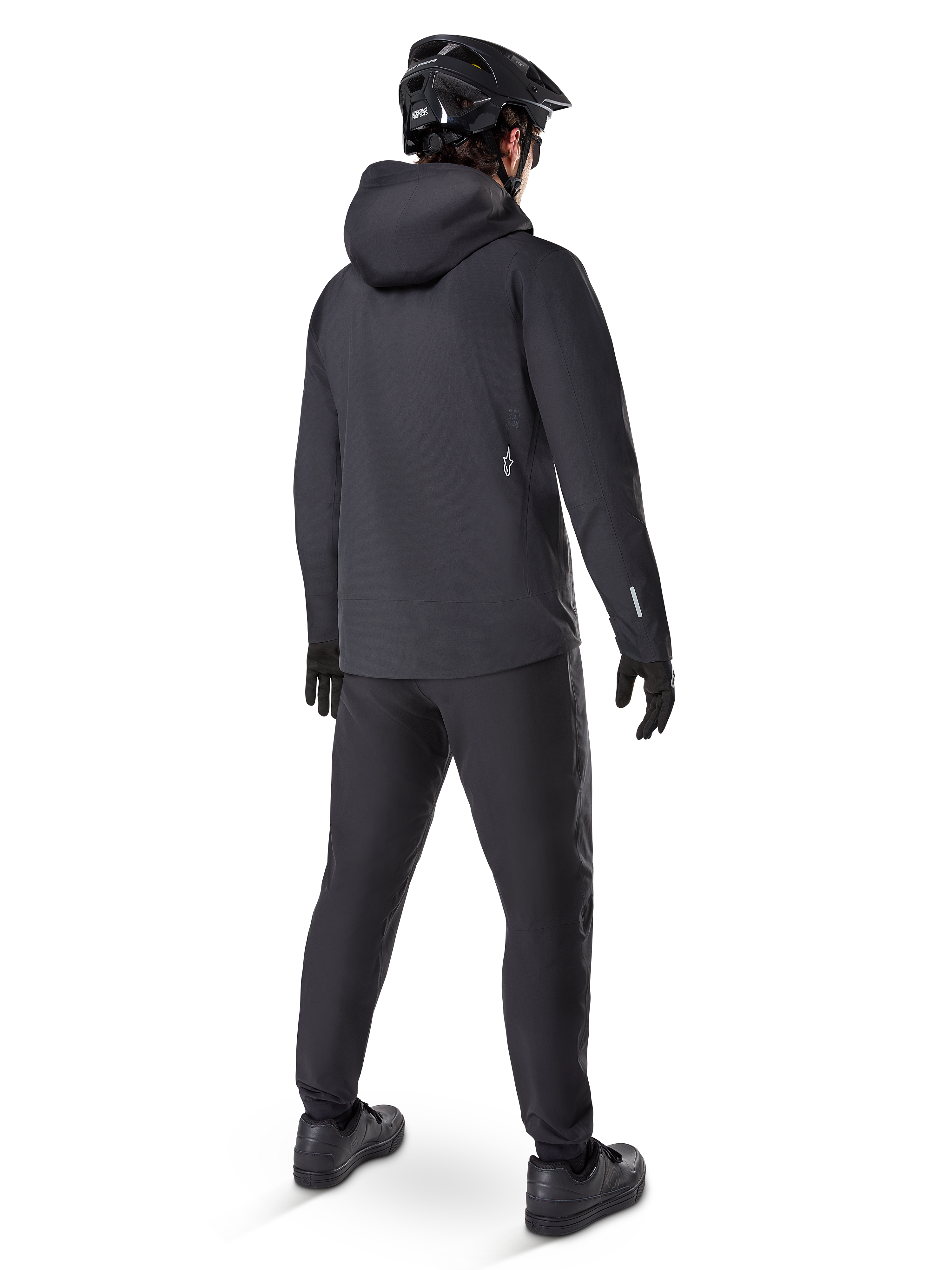 Pantaloni impermeabili Alpinestars A-Dura, pantaloni da MTB, nero, visti dal retro su un ciclista con abbigliamento da mountain bike nero completo di giacca con cappuccio e casco, abbigliamento impermeabile resistente per sentieri fangosi