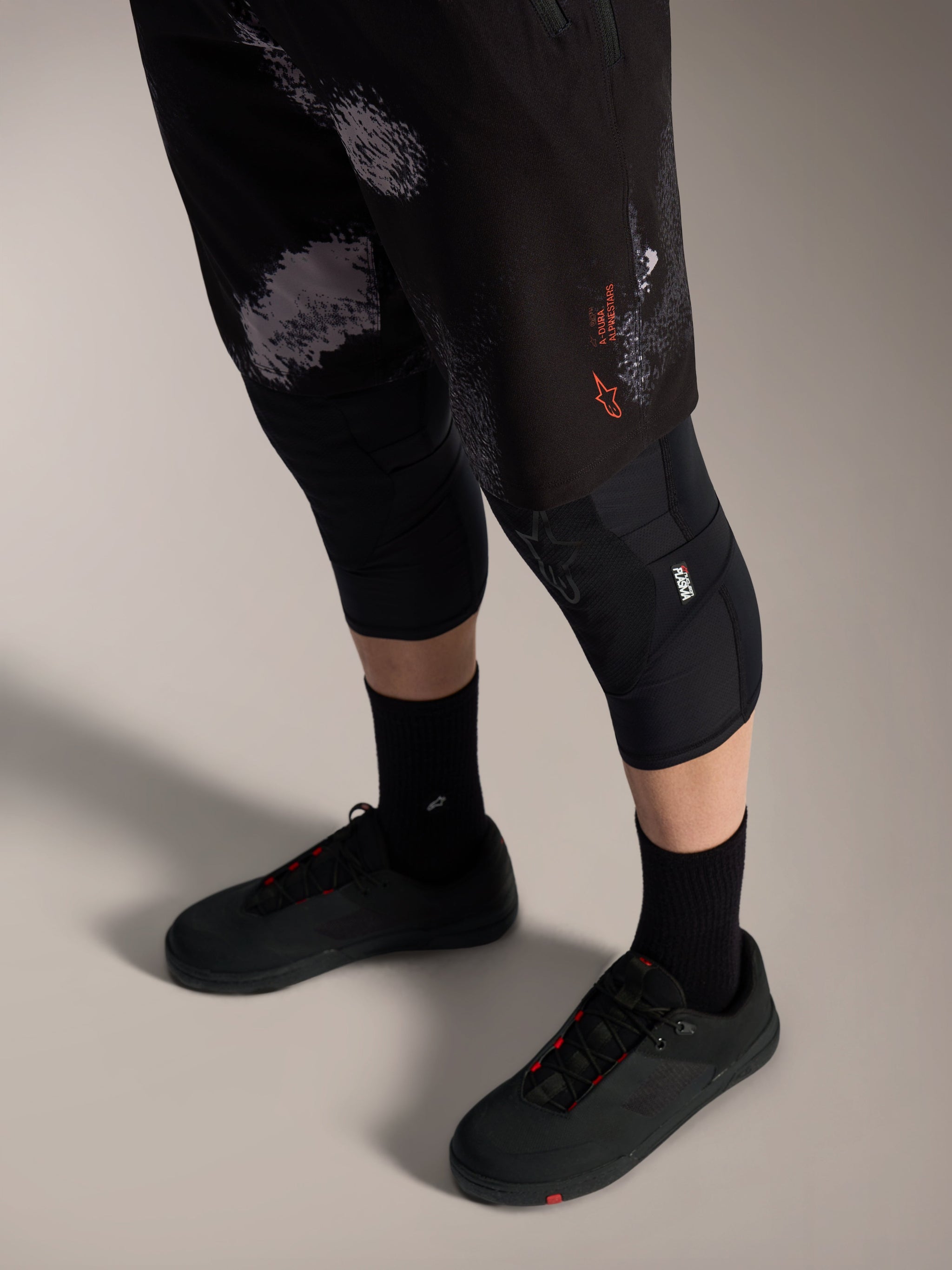 Alpinestars A-Dura Ride Shorts Black Camo, pantaloncini MTB con logo arancione, ginocchiere e scarpe tecniche.
