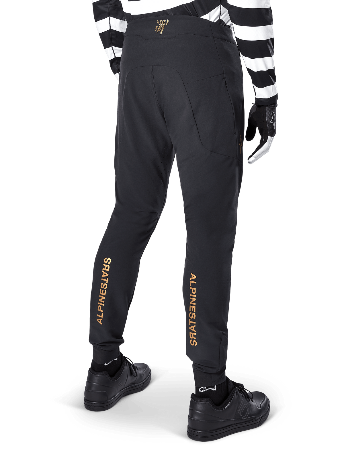 Pantaloni Alpinestars A-Aria Elite, pantaloni MTB neri, vista posteriore, con marchio Alpinestars dorato verticale sul retro delle gambe, indossati con una maglia a righe bianche e nere e scarpe nere, abbigliamento tecnico da ciclismo per mountain bike