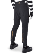 Pantaloni Alpinestars A-Aria Elite, pantaloni MTB neri, vista posteriore, con marchio Alpinestars dorato verticale sul retro delle gambe, indossati con una maglia a righe bianche e nere e scarpe nere, abbigliamento tecnico da ciclismo per mountain bike