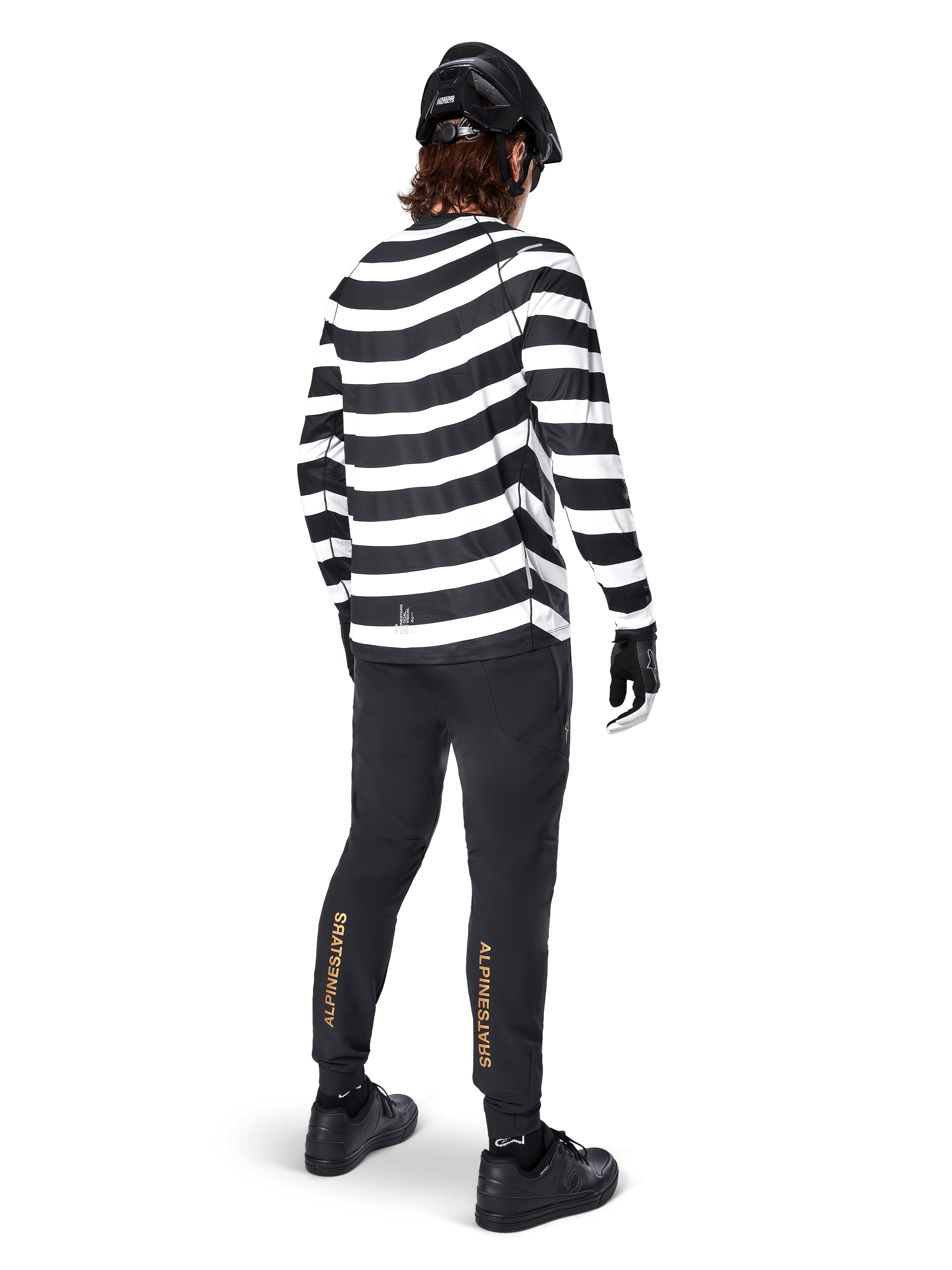 Pantaloni Alpinestars A-Aria Elite, pantaloni da MTB neri, con logo verticale dorato sui polpacci, indossati da un rider con maglia a maniche lunghe a righe bianche e nere, casco nero e scarpe nere, vista posteriore che mostra la vestibilità performante e il design pronto per il trail