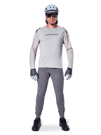 Pantaloni Alpinestars A-Aria Elite, pantaloni da mountain bike grigio scuro, indossati da un ciclista con maglia a maniche lunghe grigio chiaro, casco azzurro e scarpe nere, con perforazioni laser visibili nell'interno gamba e vestibilità slim da prestazione.