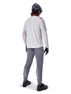 Pantaloni Alpinestars A-Aria Elite, pantaloni da ciclismo MTB, grigio scuro, vista posteriore di un rider con outfit completo, con marchio Alpinestars bianco sulla parte inferiore delle gambe, vestibilità performante con perforazioni laser, abbinati a una maglia a maniche lunghe grigio chiaro e casco da MTB grigio