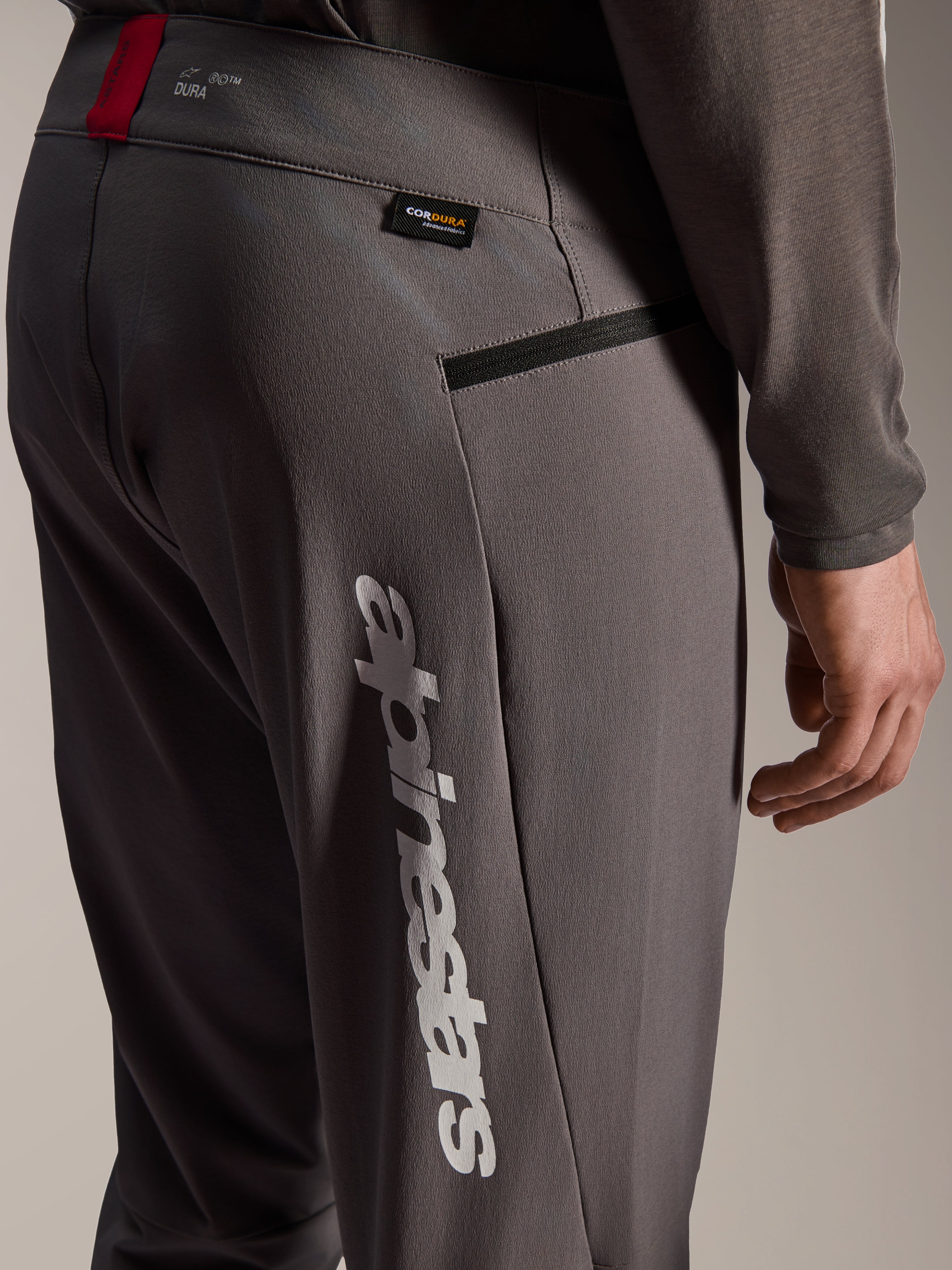 Alpinestars A-Dura Elite Pants, pantaloni da MTB grigio scuro, vista ravvicinata di fianco e coscia, con grande logo Alpinestars verticale bianco, etichetta in tessuto Cordura, tasca con zip e dettaglio rosso in vita