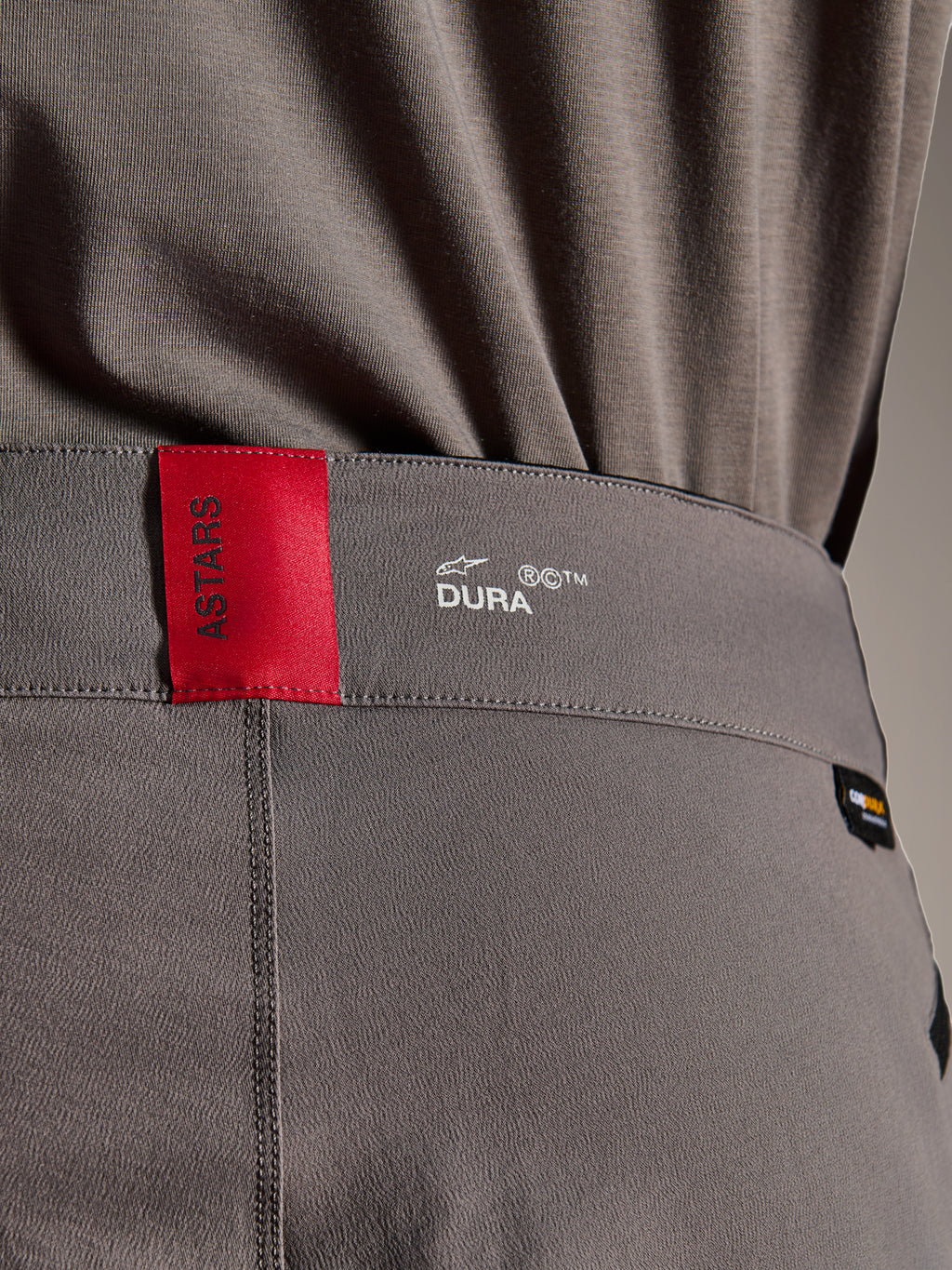 Alpinestars A-Dura Elite Pants, pantaloni da MTB, grigio scuro, primo piano della fascia in vita con etichetta rossa ASTARS e logo bianco DURA, tessuto Cordura testurizzato e dettagli di branding premium.