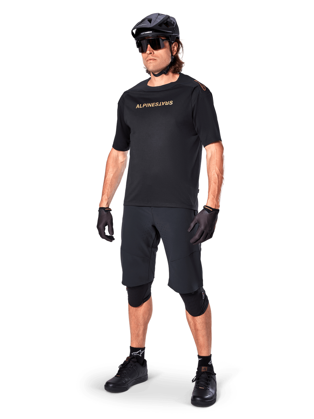 Ciclista con pantaloncini Alpinestars A-Aria Elite, pantaloncini da mountain bike neri, abbinati a una maglia nera con logo dorato, casco nero, occhiali da sole e ginocchiere, outfit tecnico completo per ciclismo fuoristrada