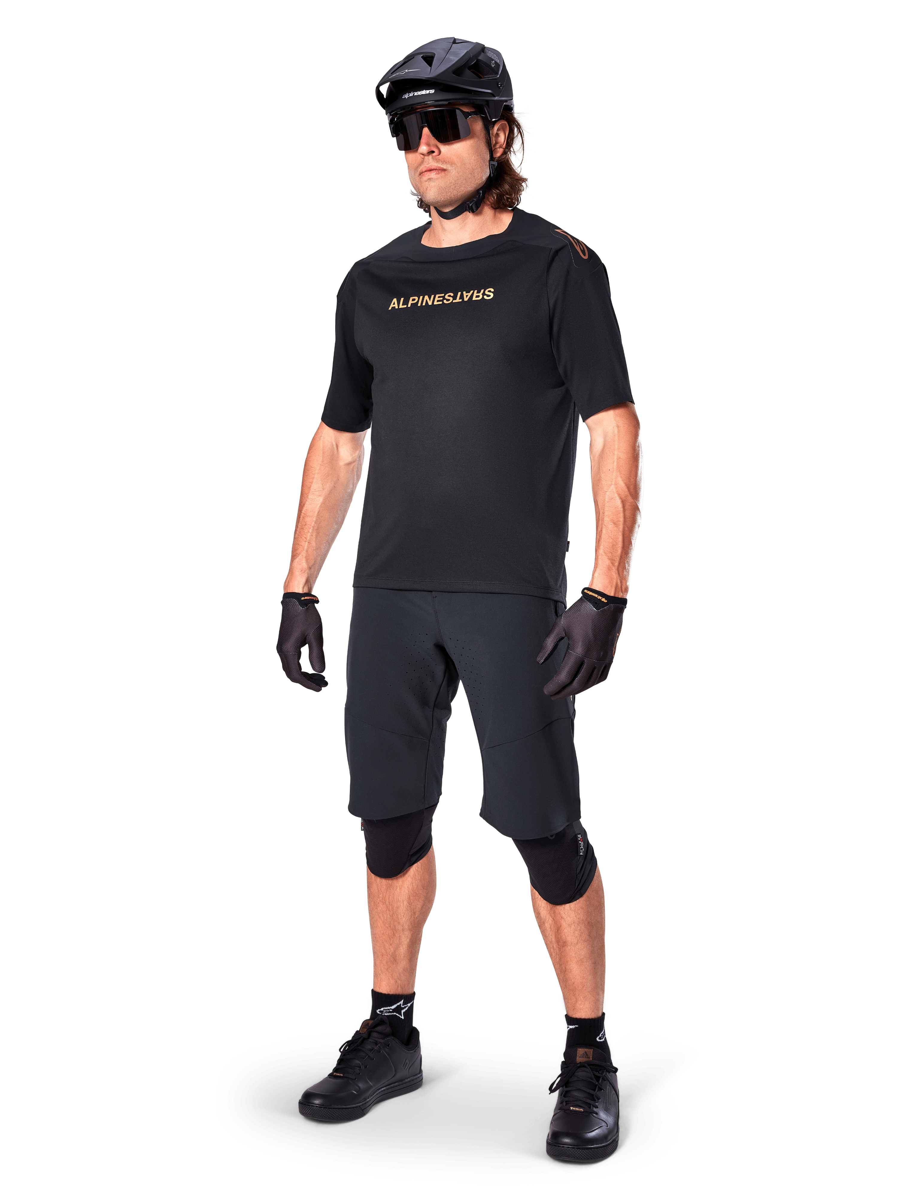 Ciclista con pantaloncini Alpinestars A-Aria Elite, pantaloncini da mountain bike neri, abbinati a una maglia nera con logo dorato, casco nero, occhiali da sole e ginocchiere, outfit tecnico completo per ciclismo fuoristrada