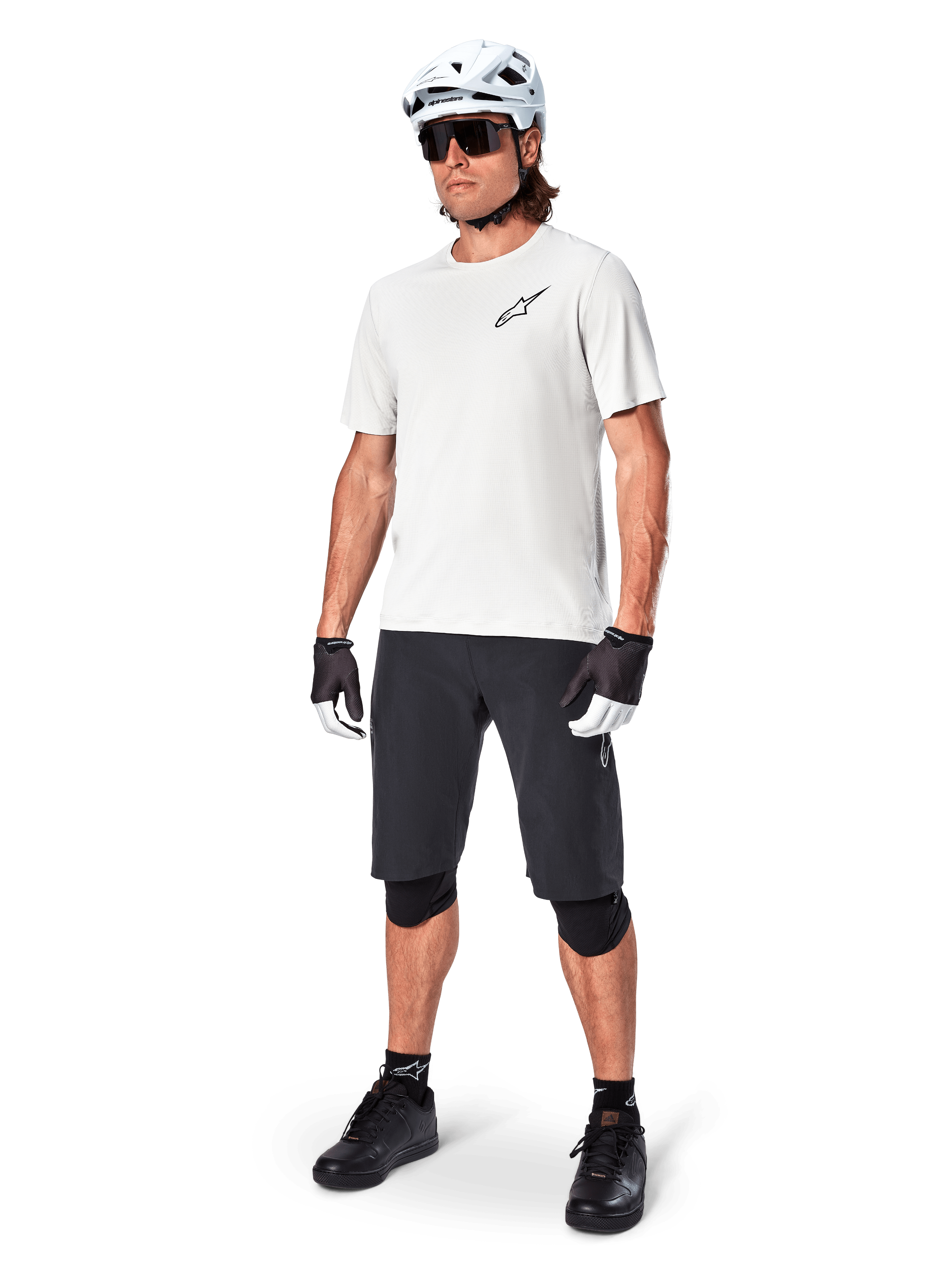 Mountain biker uomo con pantaloncini Alpinestars A-Aria, pantaloncini da ciclismo neri, abbinati a una maglia bianca a maniche corte, casco da mountain bike bianco e attrezzatura tecnica nera, outfit completo per il trail riding fuoristrada