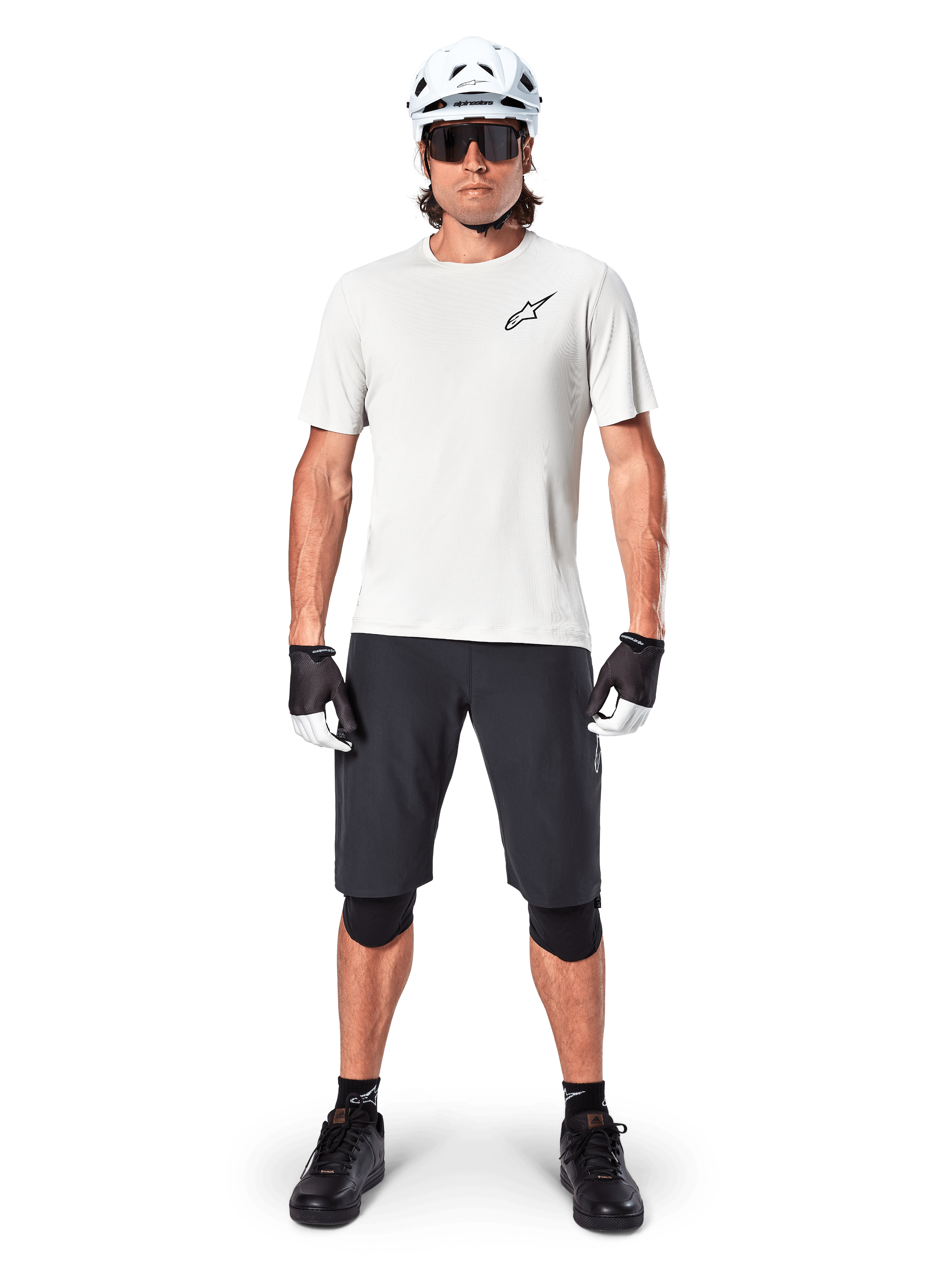 Mountain biker uomo con pantaloncini Alpinestars A-Aria neri, maglia tecnica bianca, casco bianco e protezioni per le ginocchia nere, equipaggiamento professionale da ciclismo fuoristrada in vista frontale a figura intera