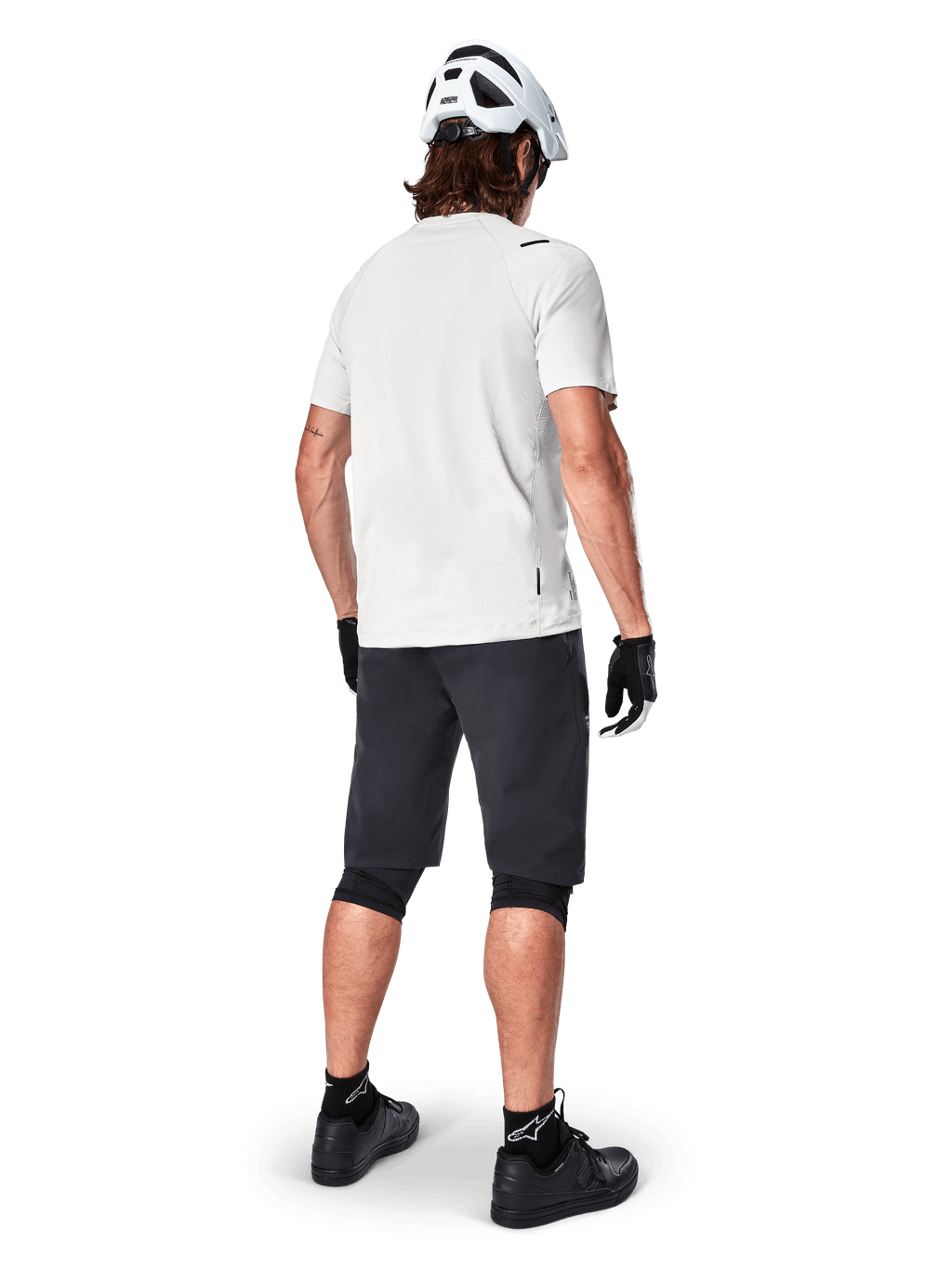 Mountain biker visto da dietro con pantaloncini Alpinestars A-Aria in nero, maglia tecnica bianca a maniche corte, casco bianco, guanti neri e scarpe da mountain bike nere, abbigliamento da ciclismo per trail riding