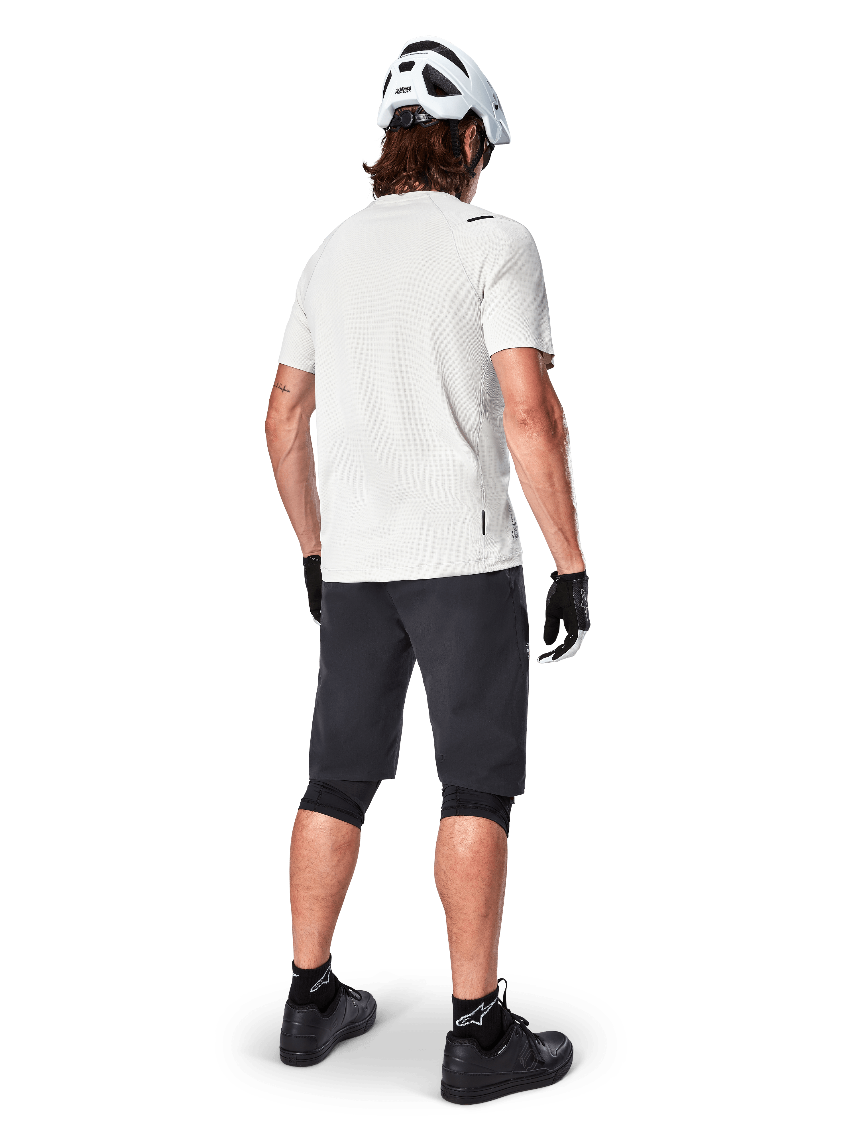 Mountain biker visto da dietro con pantaloncini Alpinestars A-Aria in nero, maglia tecnica bianca a maniche corte, casco bianco, guanti neri e scarpe da mountain bike nere, abbigliamento da ciclismo per trail riding