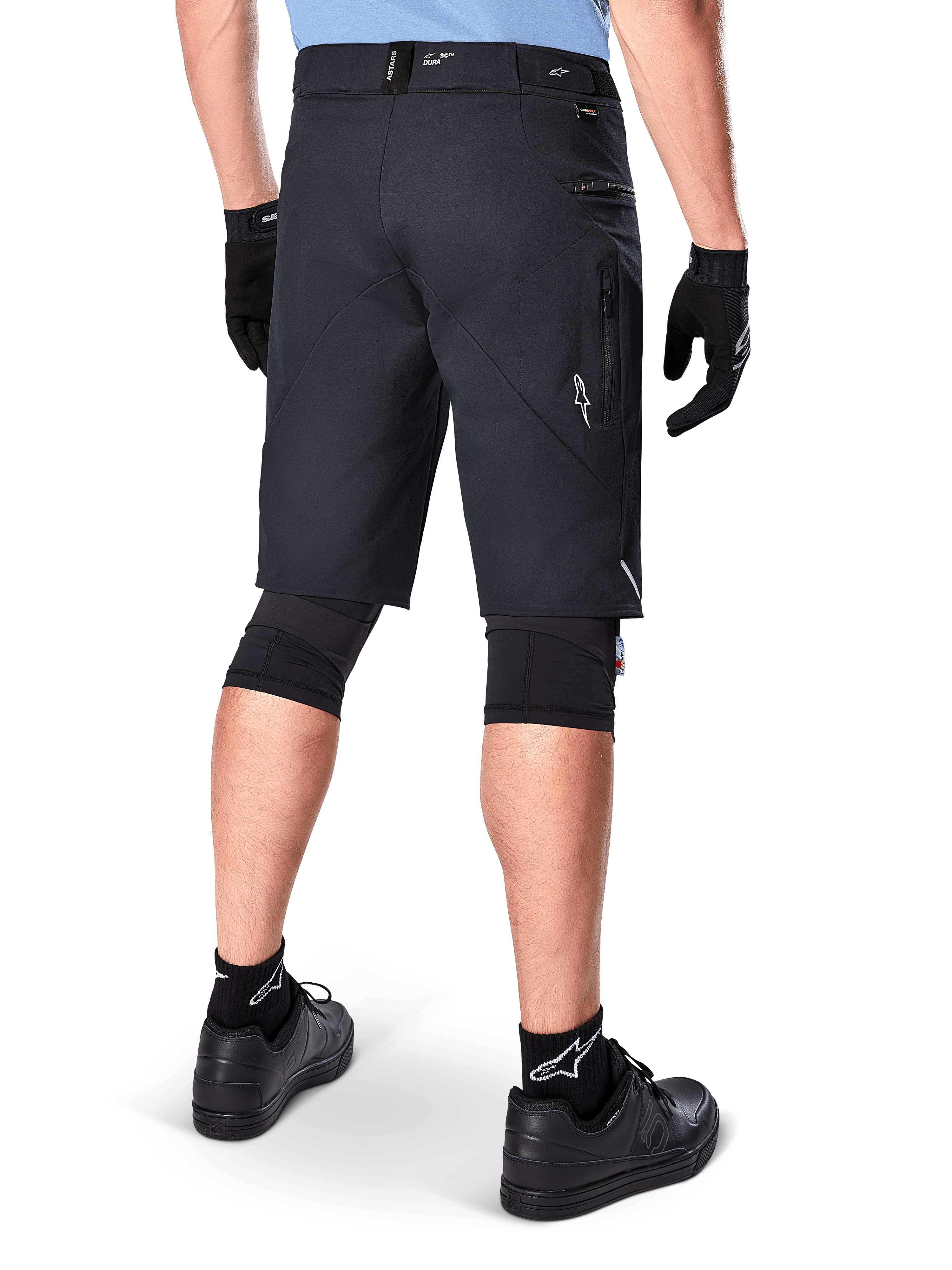 Pantaloncini Alpinestars A-Dura Elite, nero, vista posteriore di un ciclista con abbigliamento da mountain bike, incluse ginocchiere nere, guanti e calze con loghi bianchi, con tasche laterali con zip e accenti riflettenti in resistente tessuto Cordura.