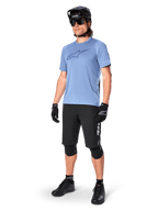Ciclista con pantaloncini Alpinestars A-Dura Elite neri, maglia a maniche corte azzurra con logo sul petto, casco nero, occhiali da sole e ginocchiere protettive, equipaggiamento completo da mountain bike per percorsi fuoristrada.