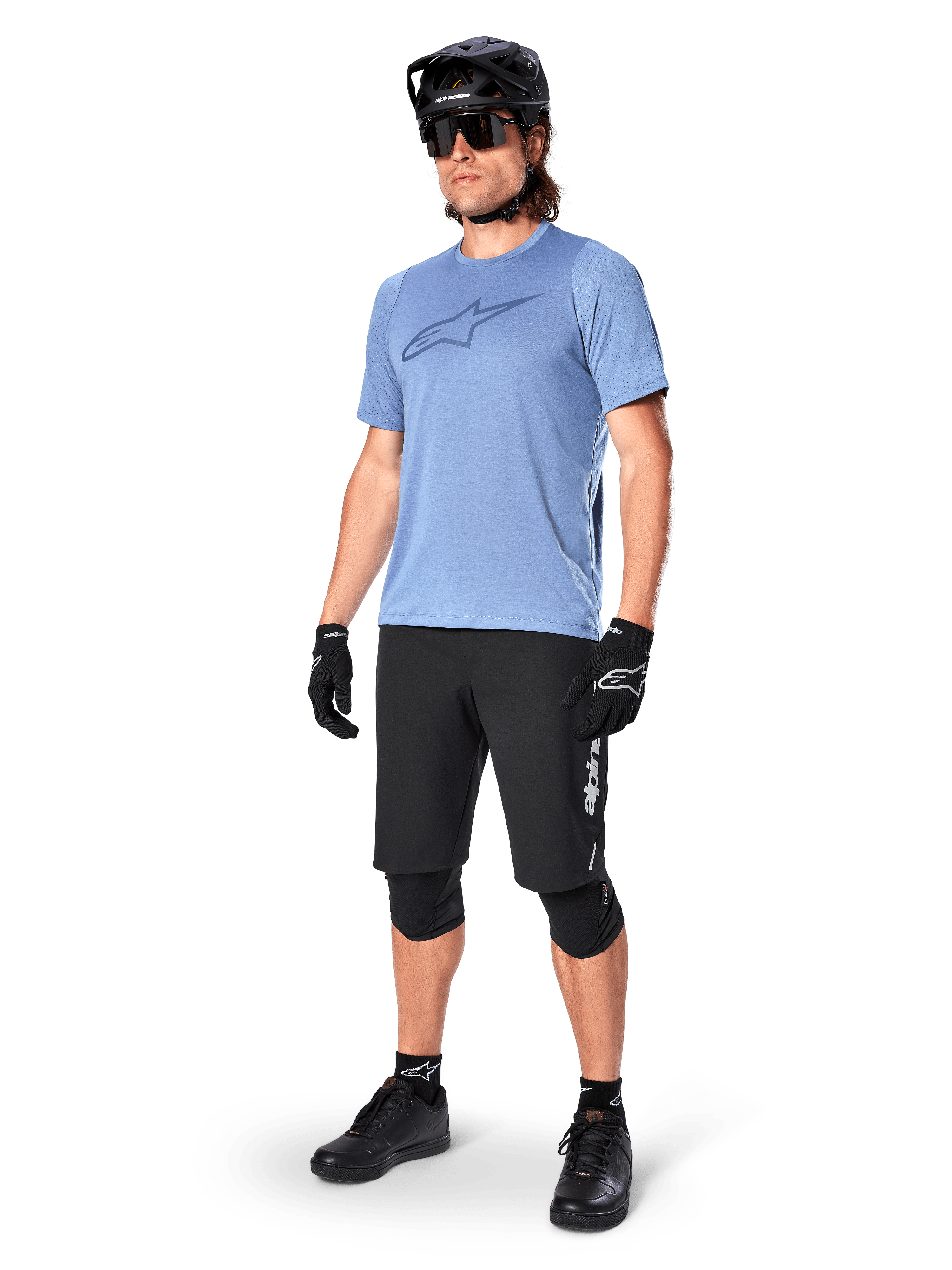 Ciclista con pantaloncini Alpinestars A-Dura Elite neri, maglia a maniche corte azzurra con logo sul petto, casco nero, occhiali da sole e ginocchiere protettive, equipaggiamento completo da mountain bike per percorsi fuoristrada.