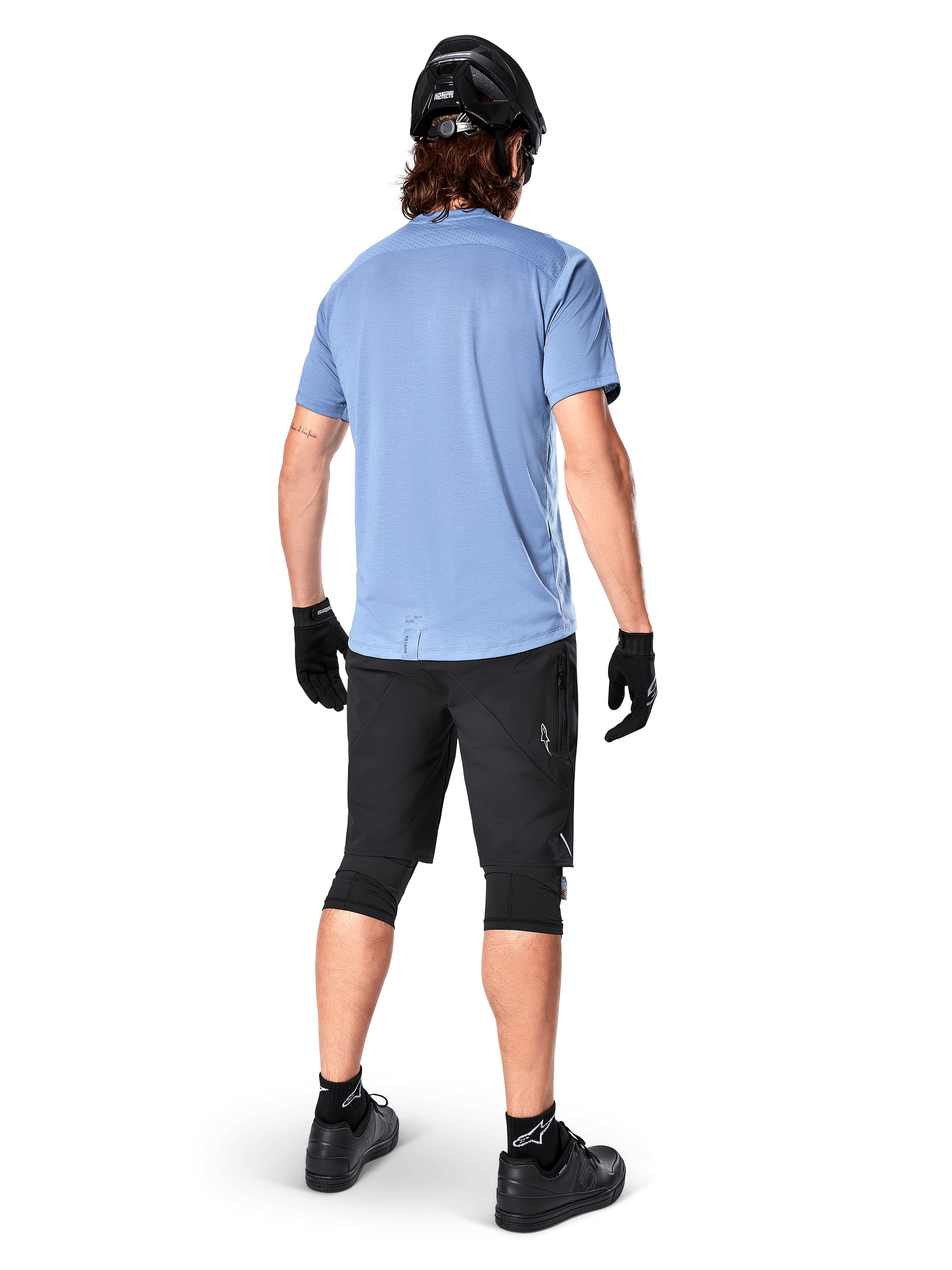 Alpinestars A-Dura Elite Shorts, pantaloncini da mountain bike, nero, vista posteriore su modello, indossati con maglia azzurra a maniche corte e attrezzatura protettiva nera, design tecnico con tasche con zip e dettagli riflettenti per il ciclismo fuoristrada