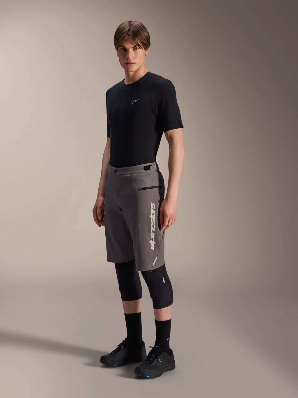 Alpinestars A-Dura Elite Shorts, pantaloncini da mountain bike grigio scuro, indossati da un modello con t-shirt nera e ginocchiere, con logo Alpinestars bianco verticale sulla gamba sinistra, tessuto tecnico Cordura per ciclismo fuoristrada
