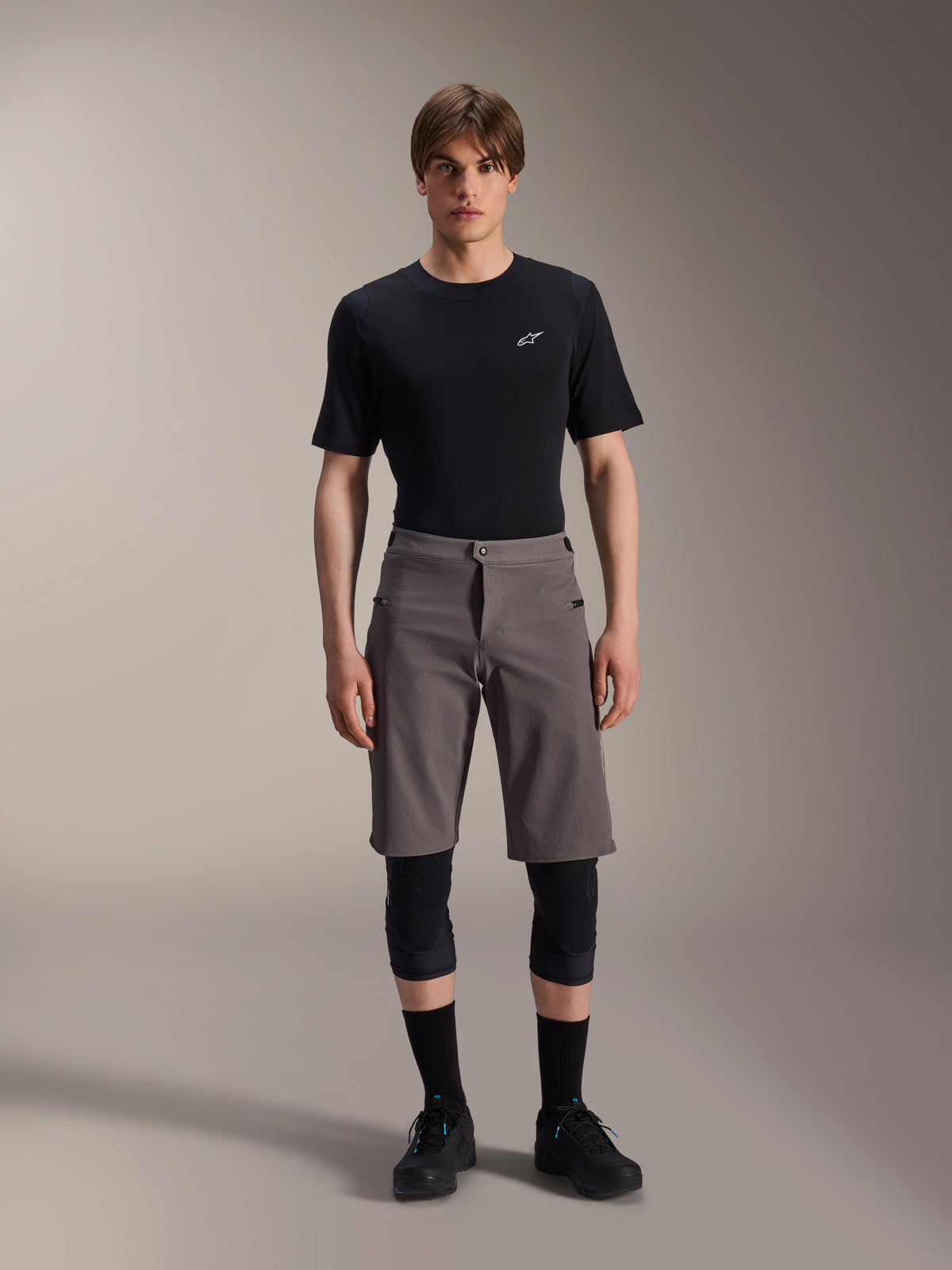 Alpinestars A-Dura Elite Shorts, pantaloncini da mountain bike grigio scuro, indossati con t-shirt nera a girocollo e ginocchiere nere, abbigliamento tecnico da ciclismo lungo fino al ginocchio con chiusura frontale a scatto e resistente tessuto Cordura.
