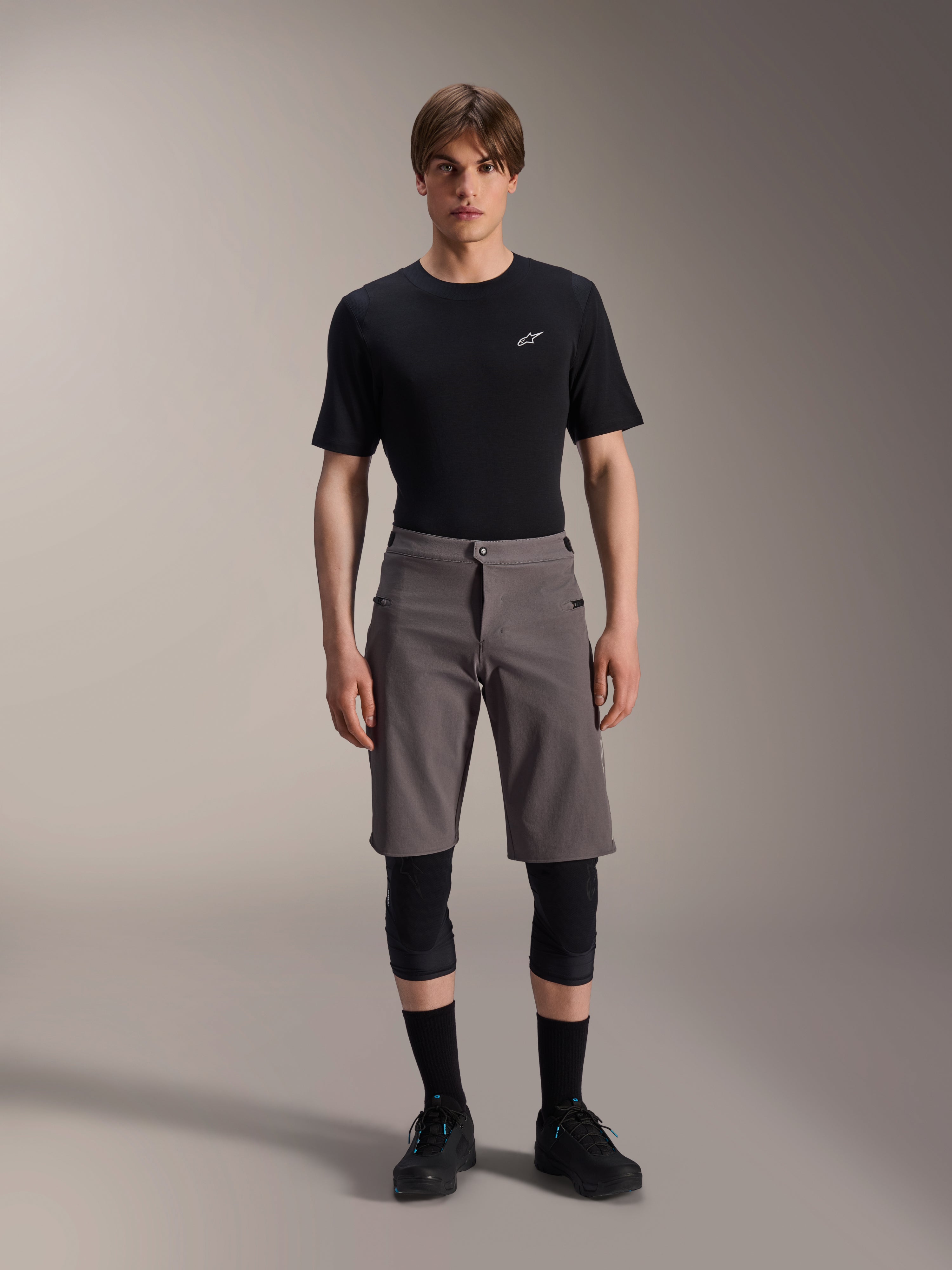 Alpinestars A-Dura Elite Shorts, pantaloncini da mountain bike grigio scuro, indossati con t-shirt nera a girocollo e ginocchiere nere, abbigliamento tecnico da ciclismo lungo fino al ginocchio con chiusura frontale a scatto e resistente tessuto Cordura.