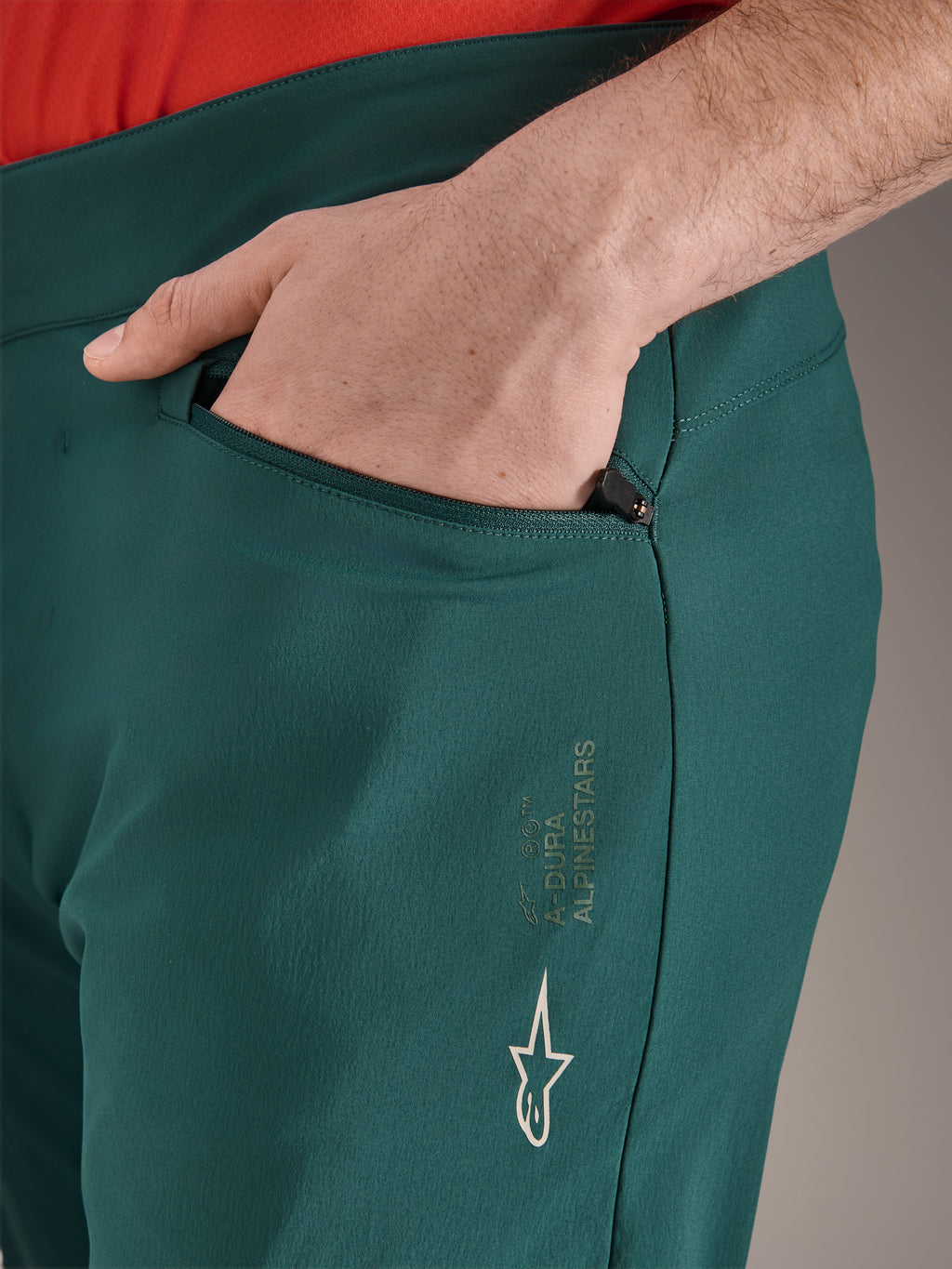 Pantaloni Alpinestars A-Dura in verde Dark Pine, primo piano della tasca con cerniera e mano, con logo Alpinestars bianco e scritta sulla coscia, pantaloni da ciclismo MTB resistenti