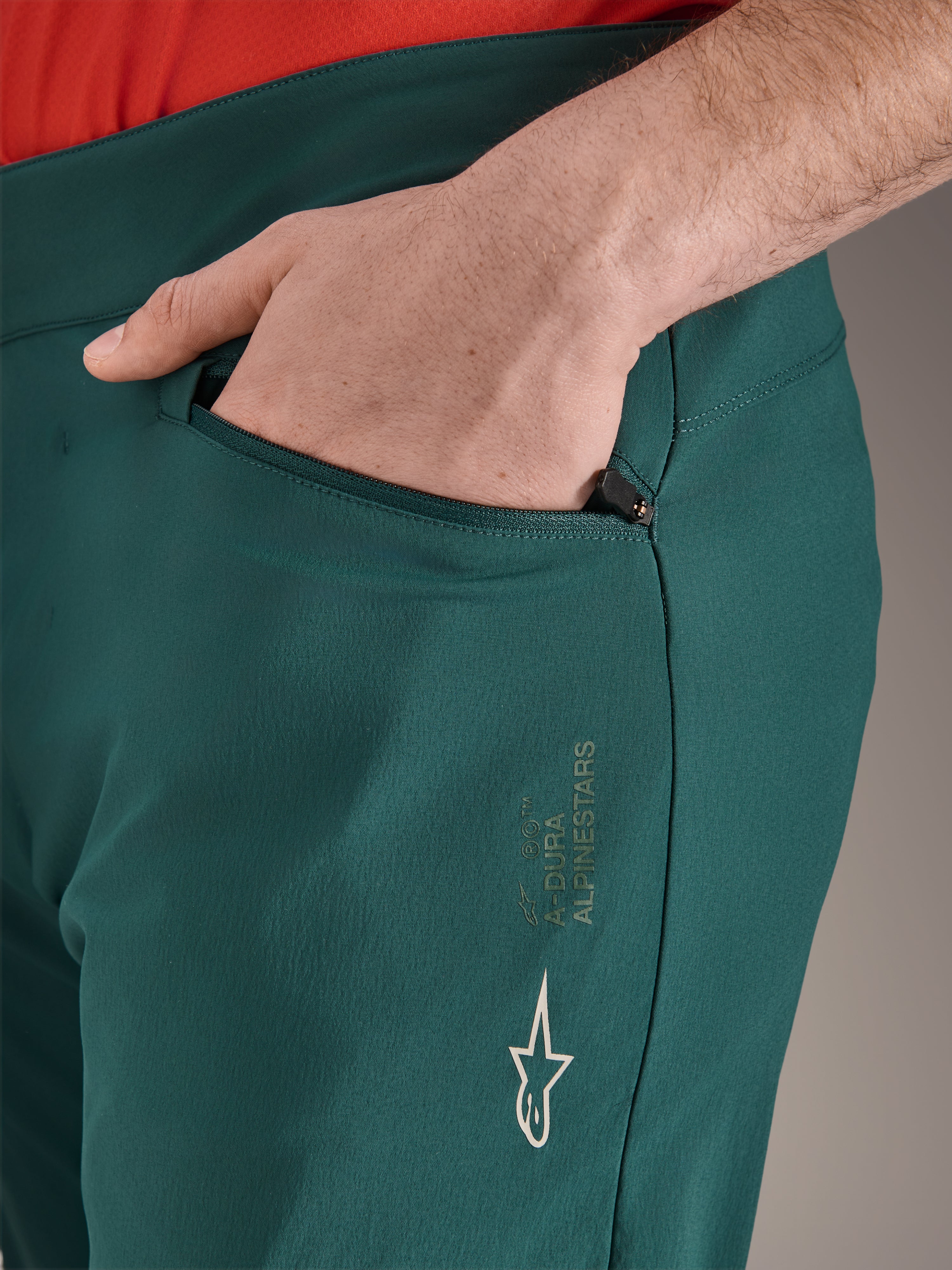 Pantaloni Alpinestars A-Dura in verde Dark Pine, primo piano della tasca con cerniera e mano, con logo Alpinestars bianco e scritta sulla coscia, pantaloni da ciclismo MTB resistenti