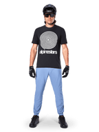 Pantaloni Alpinestars A-Dura, Infinity Blue, pantaloni da MTB indossati con una t-shirt nera con grafica a spirale, guanti neri e un casco nero, con tessuto elasticizzato resistente e polsini elasticizzati, abbigliamento tecnico da mountain bike