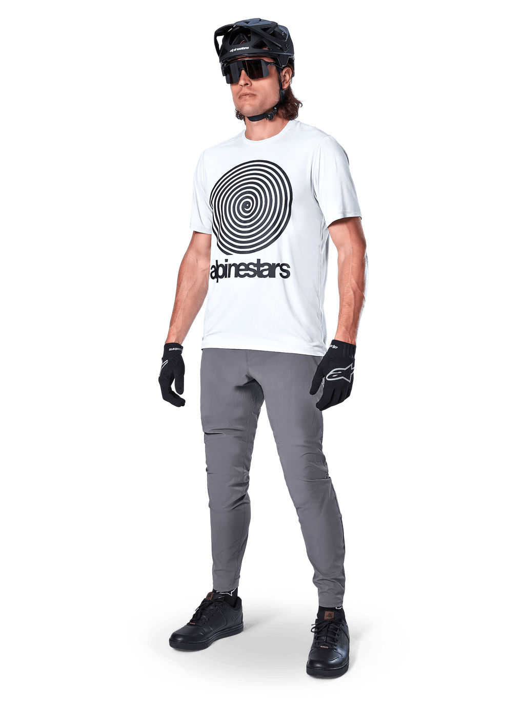 Pantaloni Alpinestars A-Dura, pantaloni da MTB, grigio scuro, indossati con t-shirt bianca con grafica a spirale, casco da MTB nero, occhiali da sole e guanti neri, tessuto resistente elasticizzato in 2 direzioni per il ciclismo fuoristrada
