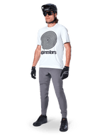 Pantaloni Alpinestars A-Dura, pantaloni da MTB, grigio scuro, indossati con t-shirt bianca con grafica a spirale, casco da MTB nero, occhiali da sole e guanti neri, tessuto resistente elasticizzato in 2 direzioni per il ciclismo fuoristrada