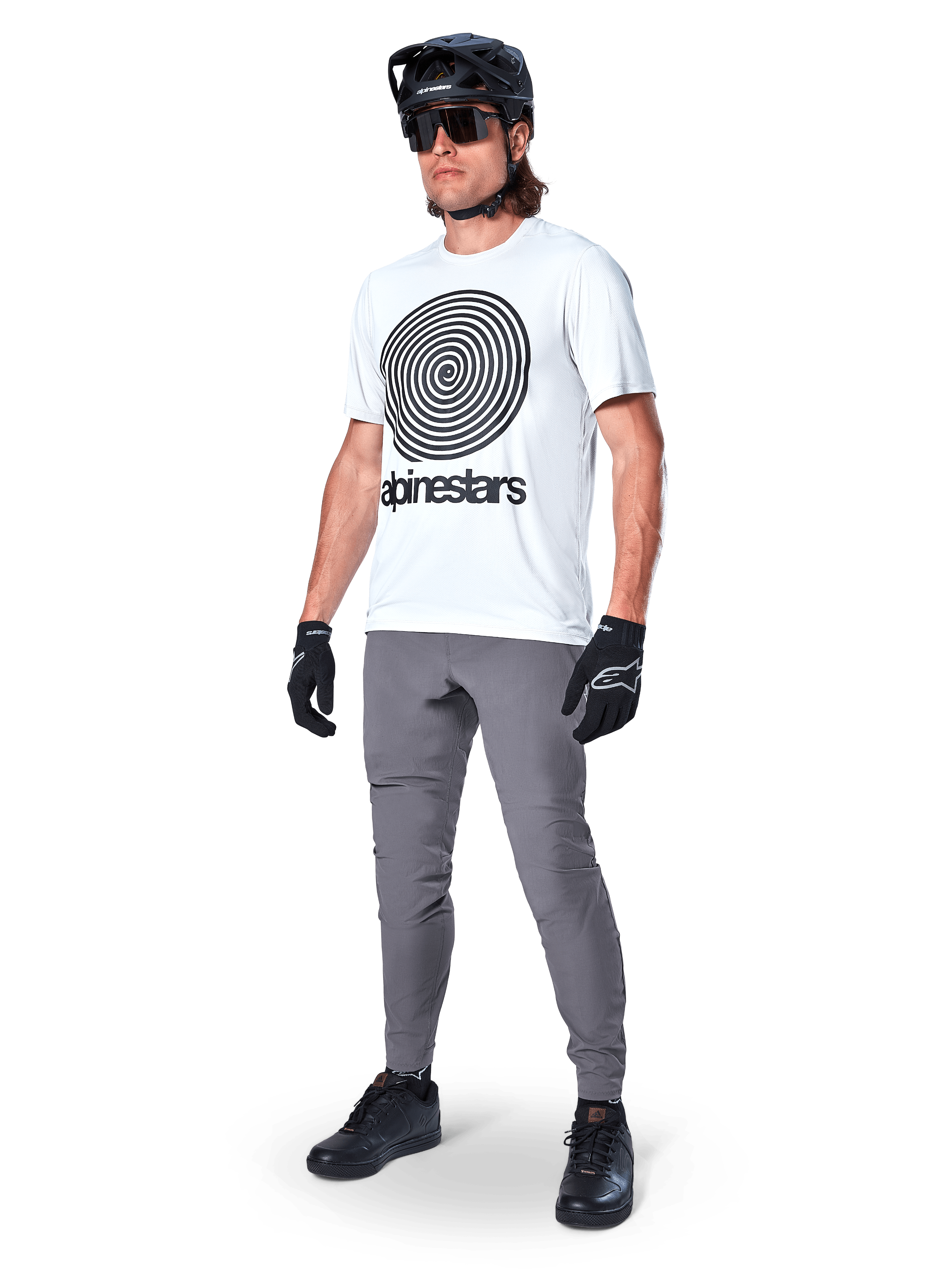 Pantaloni Alpinestars A-Dura, pantaloni da MTB, grigio scuro, indossati con t-shirt bianca con grafica a spirale, casco da MTB nero, occhiali da sole e guanti neri, tessuto resistente elasticizzato in 2 direzioni per il ciclismo fuoristrada