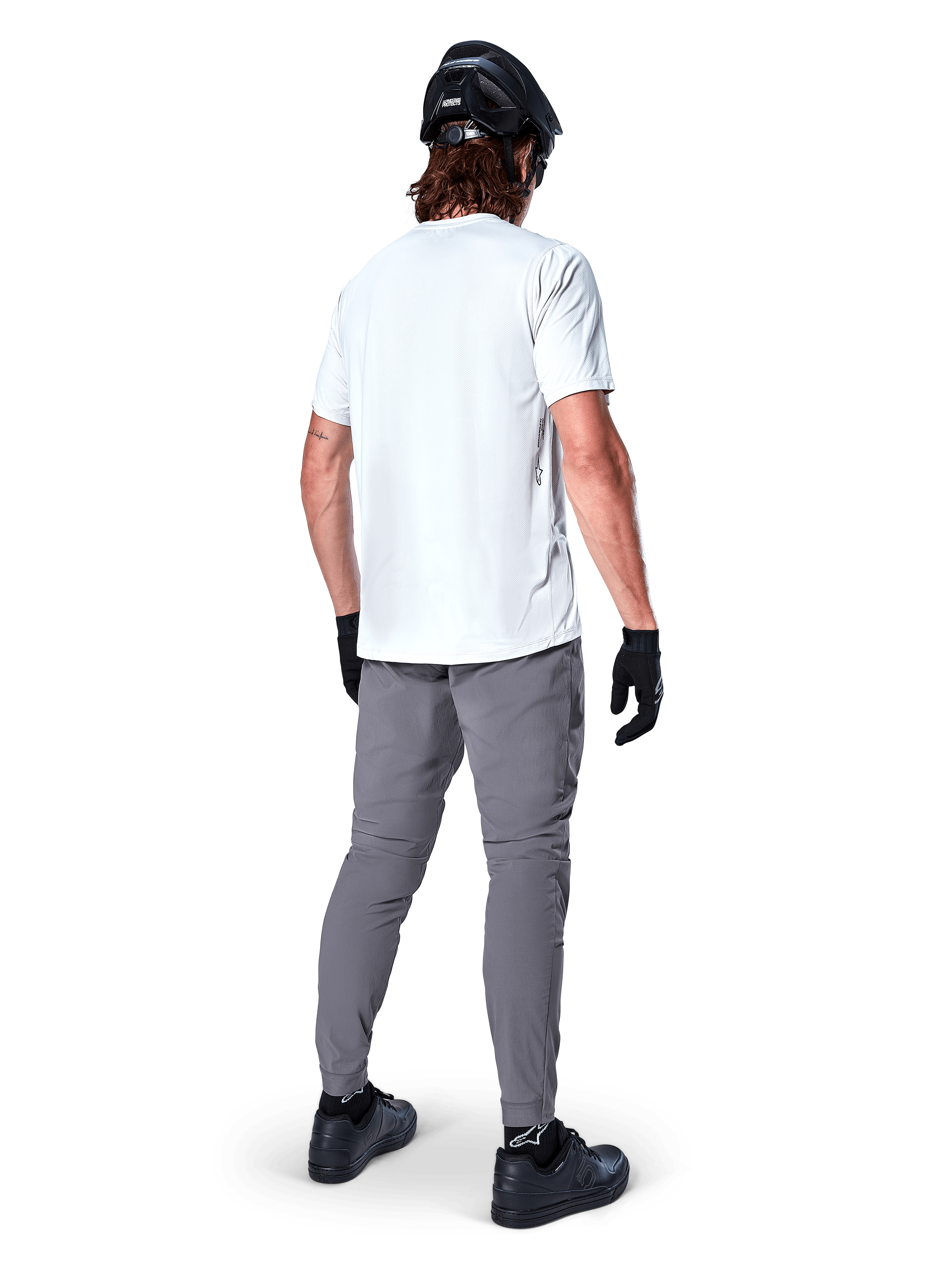 Pantaloni Alpinestars A-Dura, pantaloni da mountain bike grigio scuro visti dal retro, indossati con t-shirt bianca a maniche corte, casco da MTB nero, guanti e scarpe neri, design regular fit con polsini elasticizzati, abbigliamento resistente per ciclismo fuoristrada