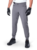 Alpinestars A-Dura Pants, pantaloni MTB grigio scuro, caratterizzati da doppia chiusura a scatto in vita e tasche con cerniera, polsini elasticizzati, indossati con guanti da ciclismo neri e sneakers nere