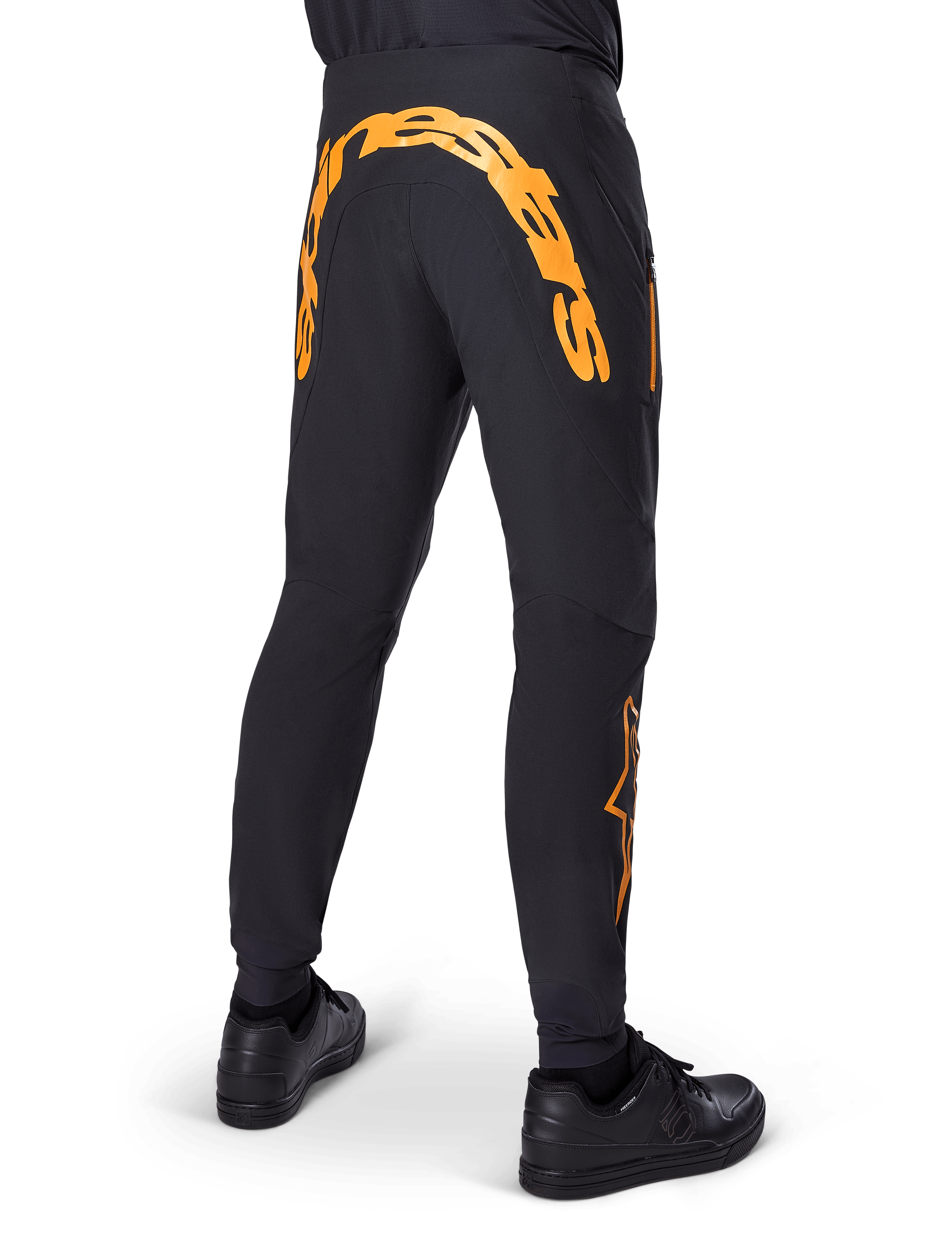 Pantaloni Alpinestars A-Supra Race, pantaloni MTB neri, vista posteriore con grande scritta Alpinestars arancione sul retro, logo arancione sulla parte inferiore della gamba, vestibilità performante con tasca con zip, indossati con scarpe nere