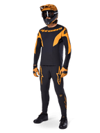 Ciclista di mountain bike con abbigliamento completo da gara, pantaloni Alpinestars A-Supra Race in nero con dettagli arancioni, maglia a maniche lunghe coordinata nera e arancione, casco arancione con maschera nera, guanti arancioni e neri e scarpe nere, design professionale per kit da gara MTB