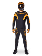 Pantaloni Alpinestars A-Supra Race, pantaloni da gara MTB, nero e arancione, indossati da un pilota con un kit da gara completo composto da maglia nera e arancione, casco arancione e guanti coordinati, con grandi loghi a stella arancioni sulle gambe