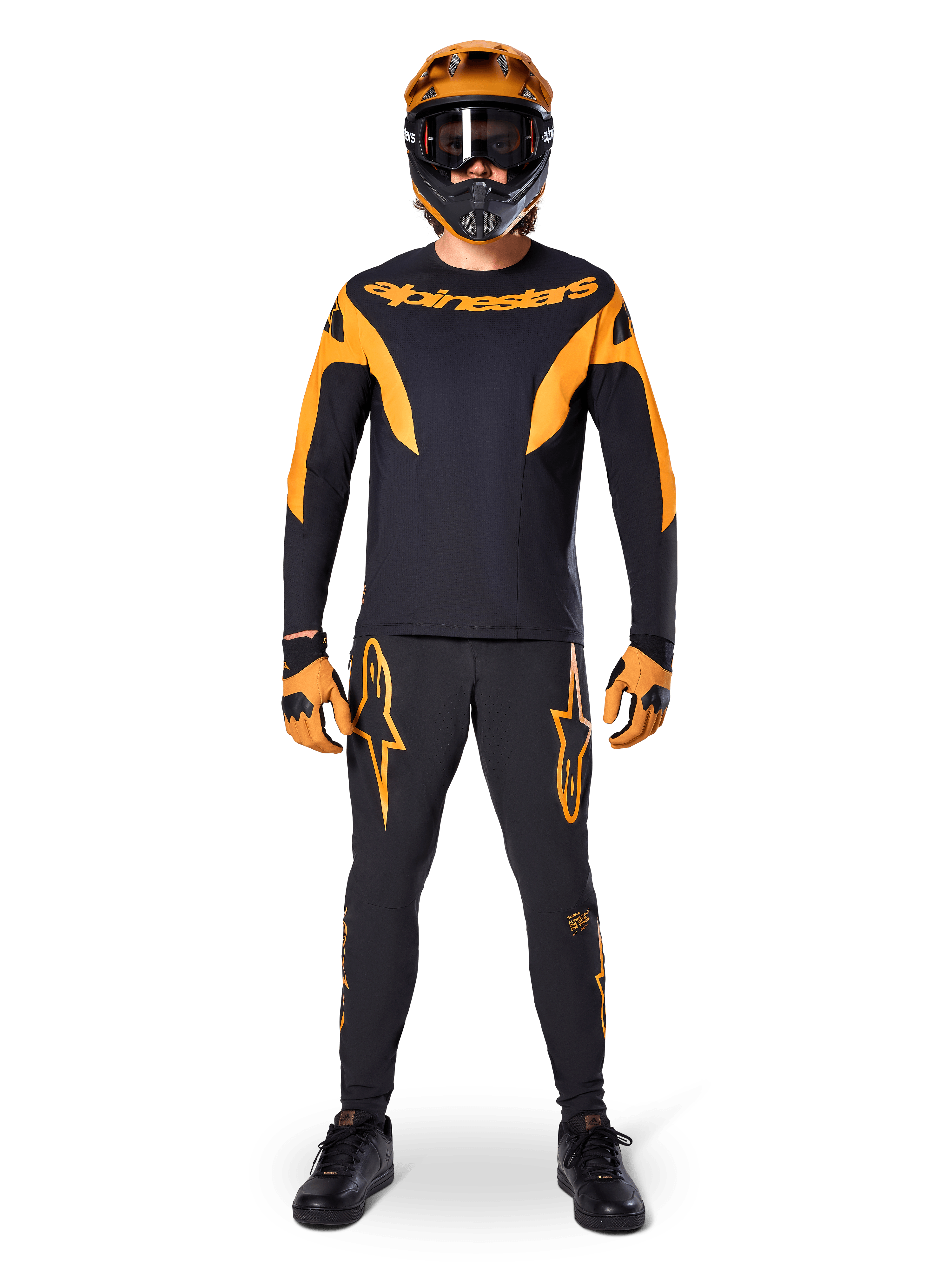 Pantaloni Alpinestars A-Supra Race, pantaloni da gara MTB, nero e arancione, indossati da un pilota con un kit da gara completo composto da maglia nera e arancione, casco arancione e guanti coordinati, con grandi loghi a stella arancioni sulle gambe