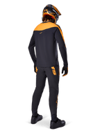 Alpinestars A-Supra Race Pants, pantaloni da gara MTB, neri con loghi arancioni, vista posteriore su un rider che indossa maglia, casco e guanti coordinati nero e arancione, evidenziando il wordmark ispirato al MX sul carré posteriore e la vestibilità performante per trail tecnici.