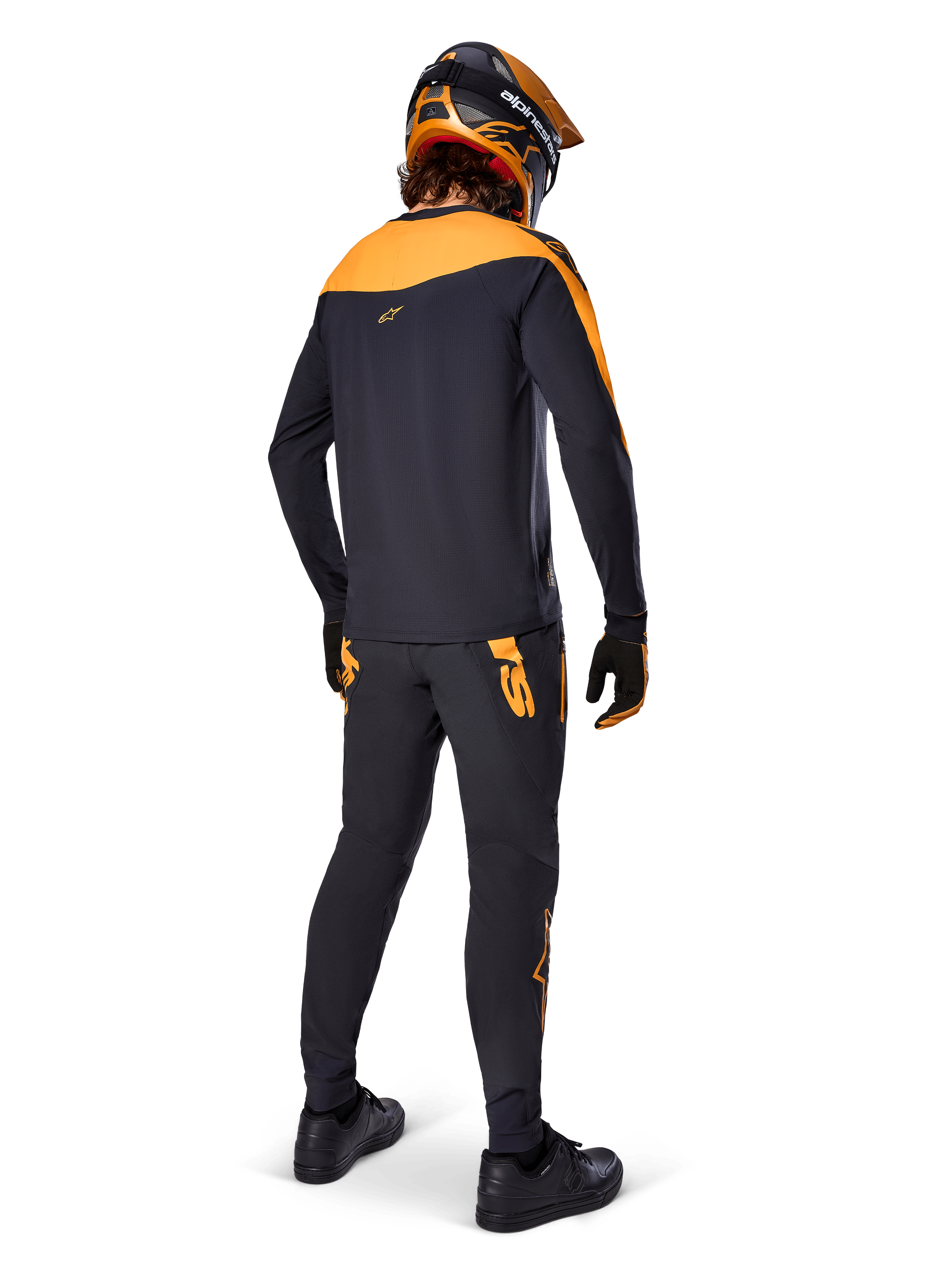 Alpinestars A-Supra Race Pants, pantaloni da gara MTB, neri con loghi arancioni, vista posteriore su un rider che indossa maglia, casco e guanti coordinati nero e arancione, evidenziando il wordmark ispirato al MX sul carré posteriore e la vestibilità performante per trail tecnici.