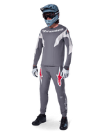 Alpinestars pantaloni A-Supra Race in grigio scuro, con vistosi loghi rossi e bianchi sulle cosce, indossati da un rider come parte di un kit completo da mountain bike con maglia grigia coordinata, casco integrale grigio-blu e guanti grigio chiaro, abbigliamento ad alte prestazioni per gare MTB