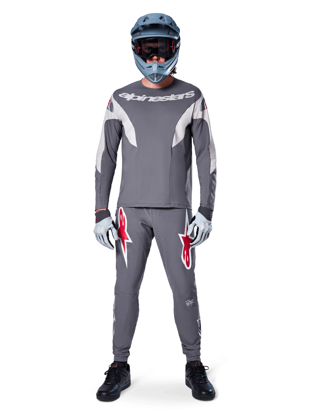 Rider MTB con pantaloni Alpinestars A-Supra Race in grigio scuro, caratterizzati da vivaci loghi rossi sulle cosce, abbinati a maglia grigia coordinata, casco integrale blu e scarpe nere per un kit completo da gara off-road.