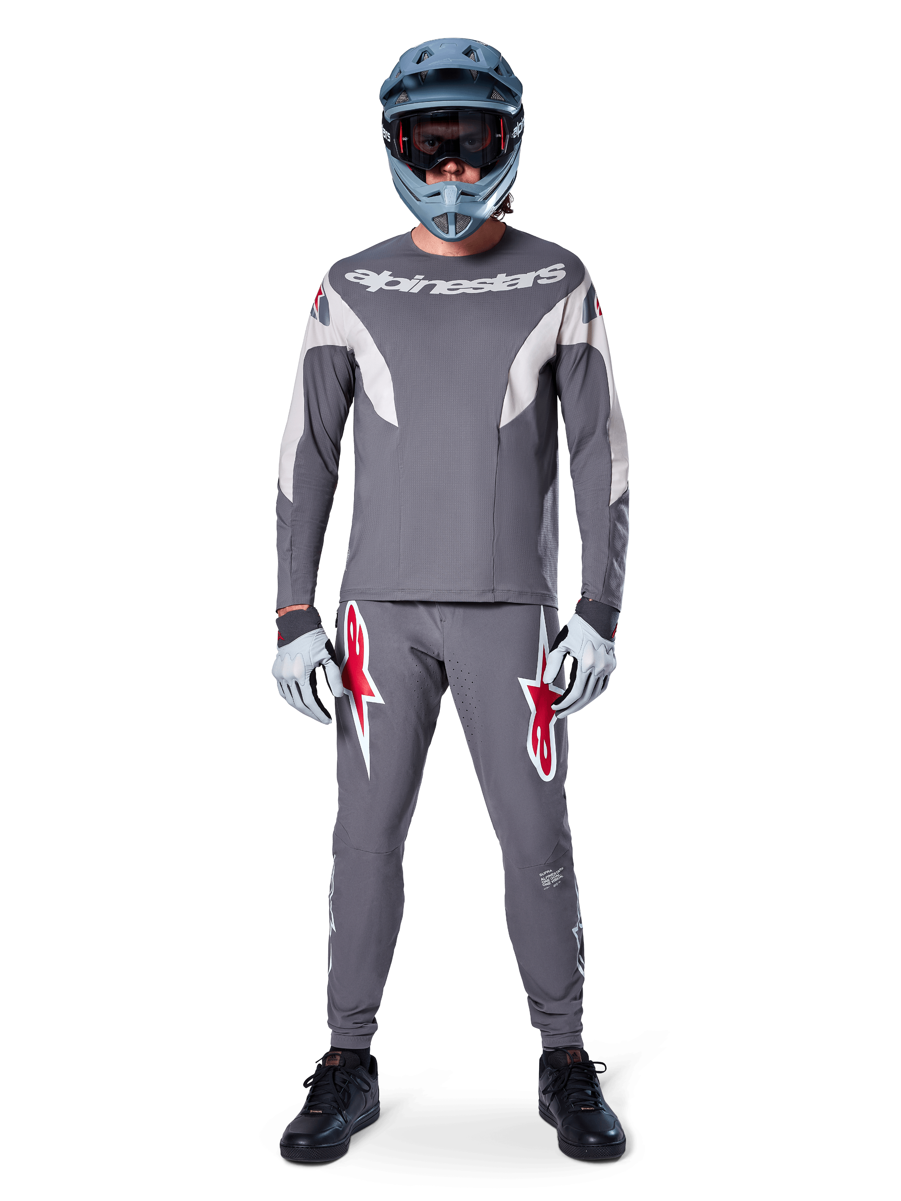 Rider MTB con pantaloni Alpinestars A-Supra Race in grigio scuro, caratterizzati da vivaci loghi rossi sulle cosce, abbinati a maglia grigia coordinata, casco integrale blu e scarpe nere per un kit completo da gara off-road.