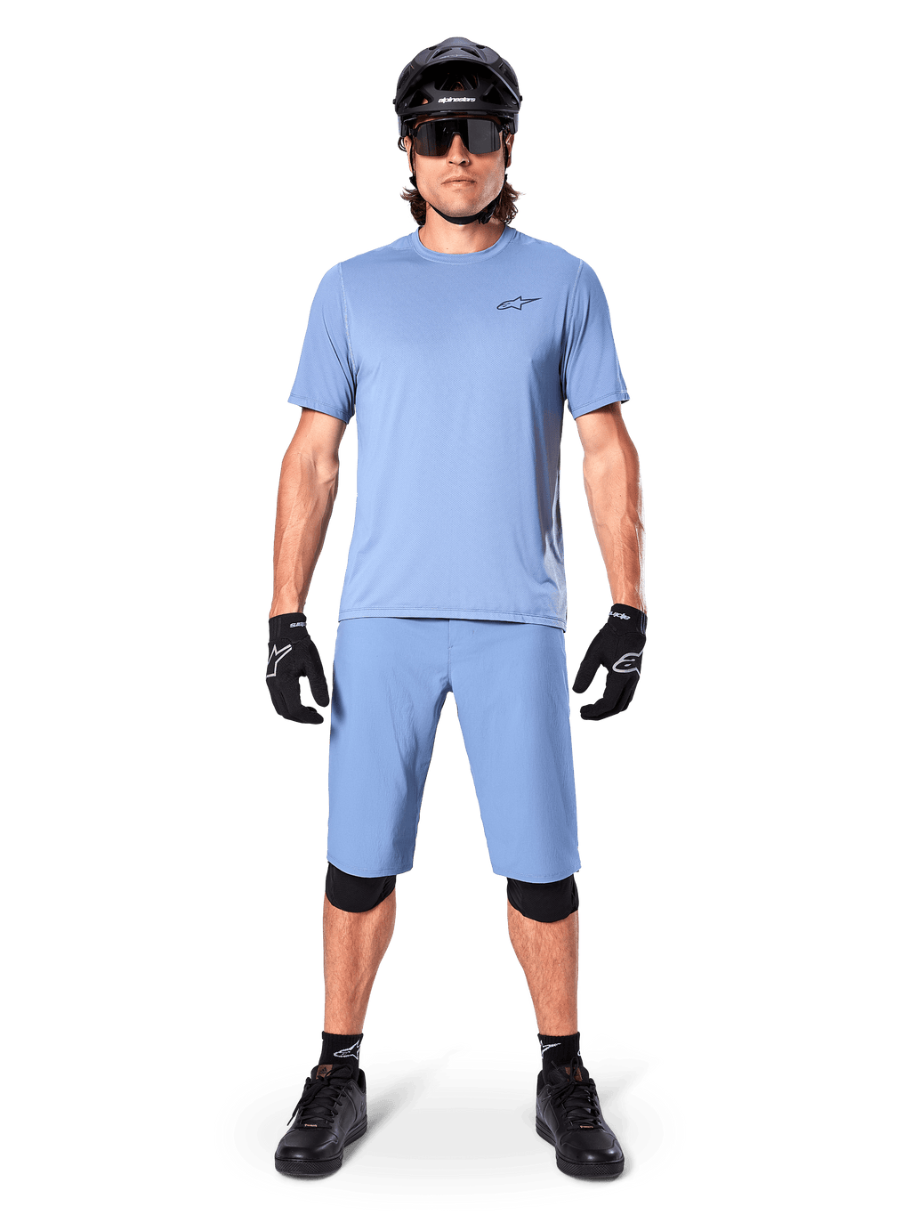 Modello con pantaloncini Alpinestars A-Dura in Infinity Blue, maglia azzurra coordinata, casco da mountain bike nero, occhiali da sole neri, guanti e ginocchiere neri, outfit completo da MTB per trail riding