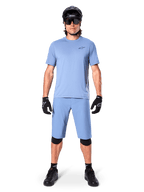 Modello con pantaloncini Alpinestars A-Dura in Infinity Blue, maglia azzurra coordinata, casco da mountain bike nero, occhiali da sole neri, guanti e ginocchiere neri, outfit completo da MTB per trail riding