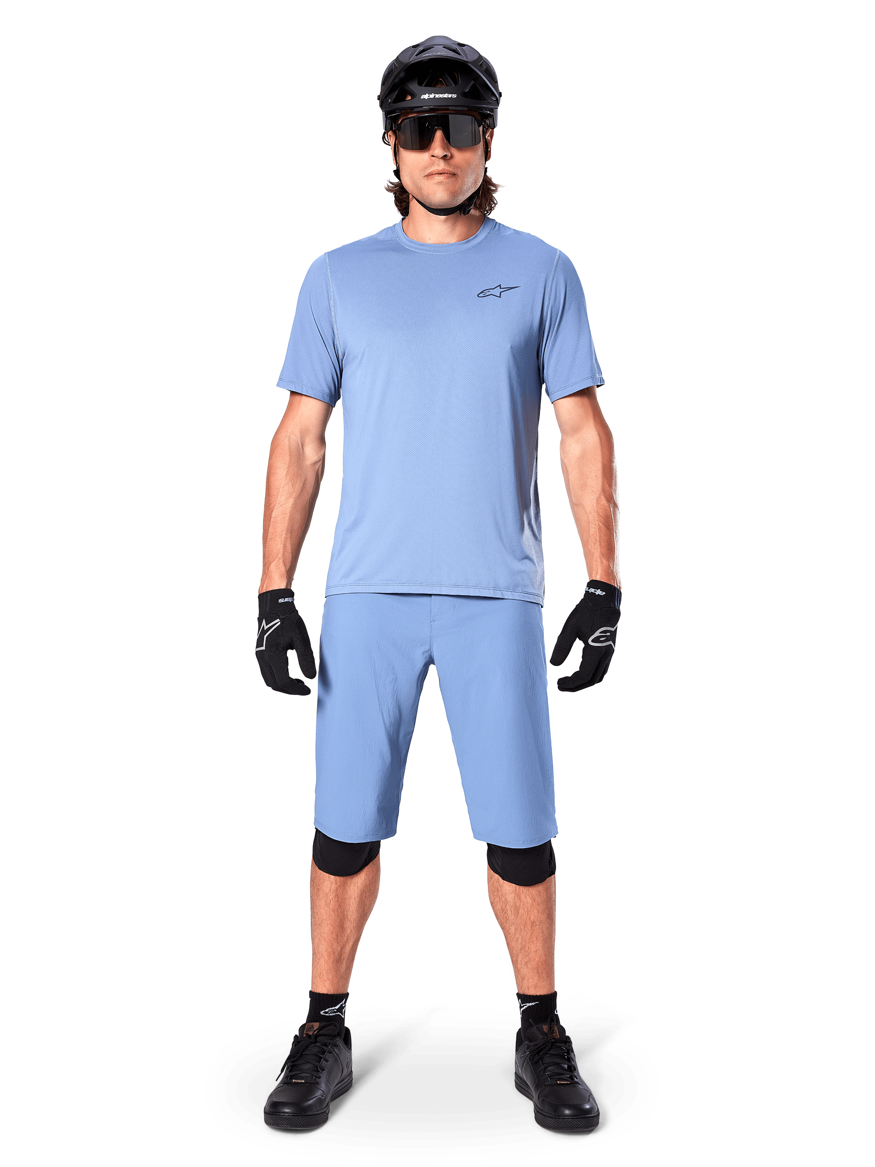 Modello con pantaloncini Alpinestars A-Dura in Infinity Blue, maglia azzurra coordinata, casco da mountain bike nero, occhiali da sole neri, guanti e ginocchiere neri, outfit completo da MTB per trail riding