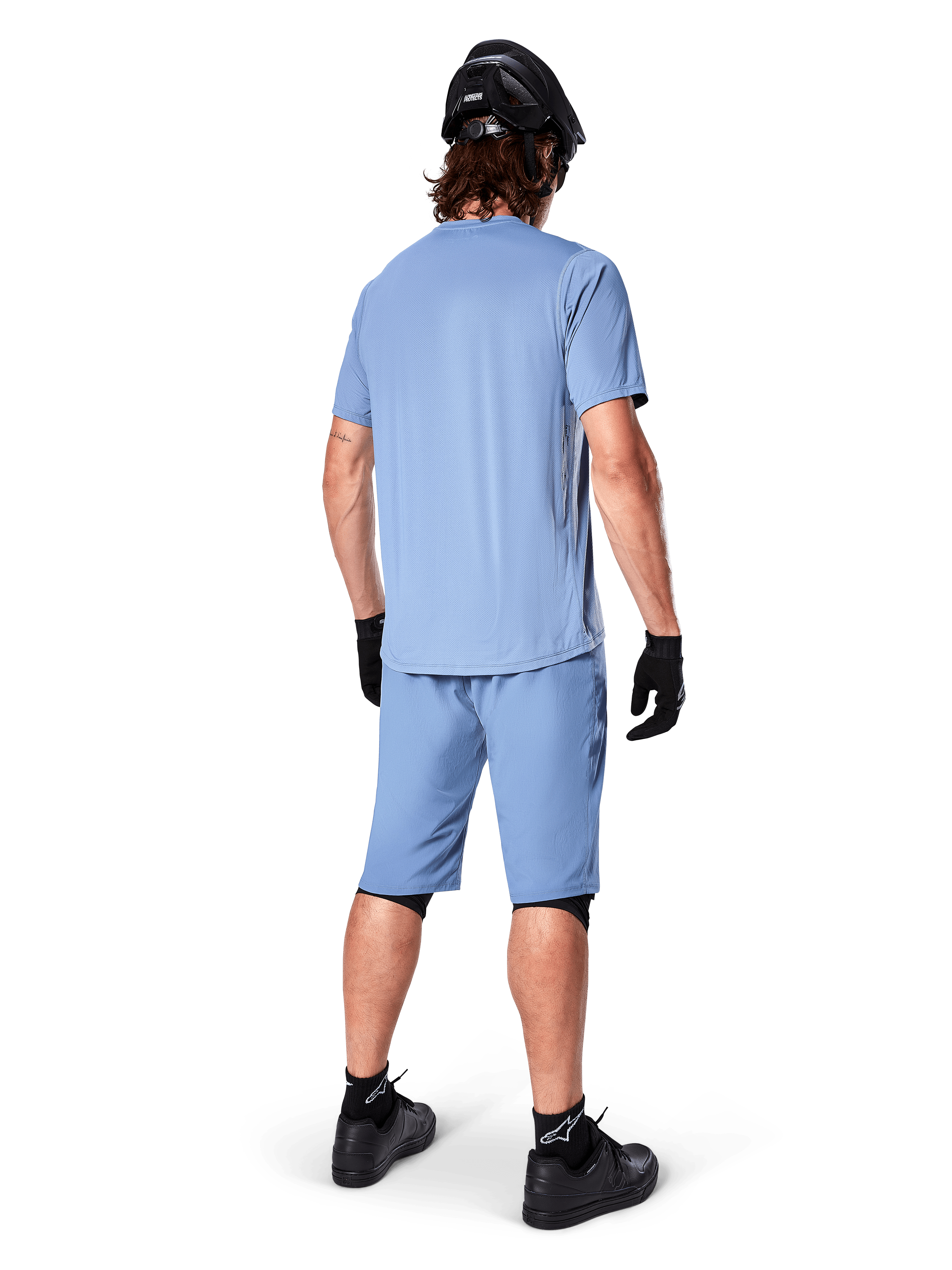 Vista posteriore di un mountain biker con pantaloncini Alpinestars A-Dura in Infinity Blue, maglia a maniche corte coordinata, casco, guanti e scarpe neri, abbigliamento tecnico coordinato per trail riding in MTB