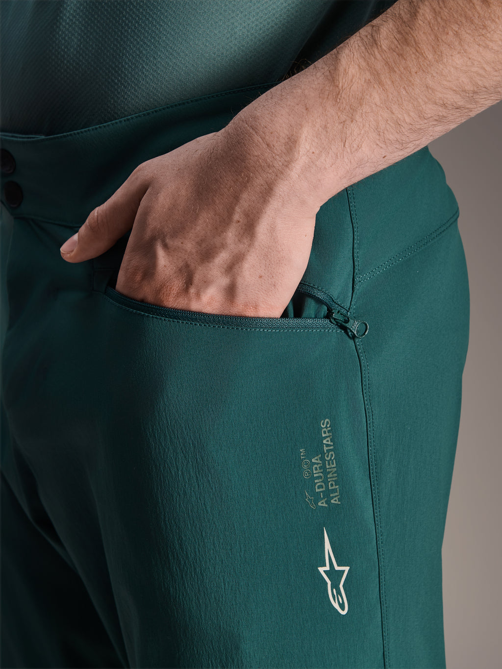 Alpinestars Shorts A-Dura in Dark Pine, vista ravvicinata di una mano nella tasca con zip foderata in rete, con logo a stella Alpinestars bianco e marchio A-Dura, costruzione in tessuto resistente per mountain bike