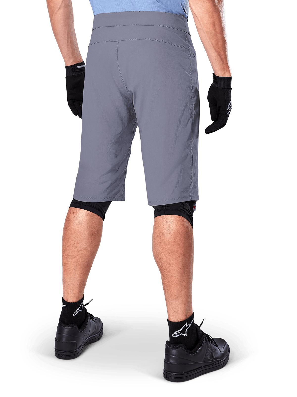 Alpinestars A-Dura Shorts, pantaloncini da mountain bike grigio scuro, vista posteriore su un ciclista, indossati con guanti neri, protezioni ginocchia nere e calze nere con logo Alpinestars, costruzione resistente per il ciclismo fuoristrada