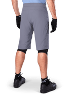 Alpinestars A-Dura Shorts, pantaloncini da mountain bike grigio scuro, vista posteriore su un ciclista, indossati con guanti neri, protezioni ginocchia nere e calze nere con logo Alpinestars, costruzione resistente per il ciclismo fuoristrada