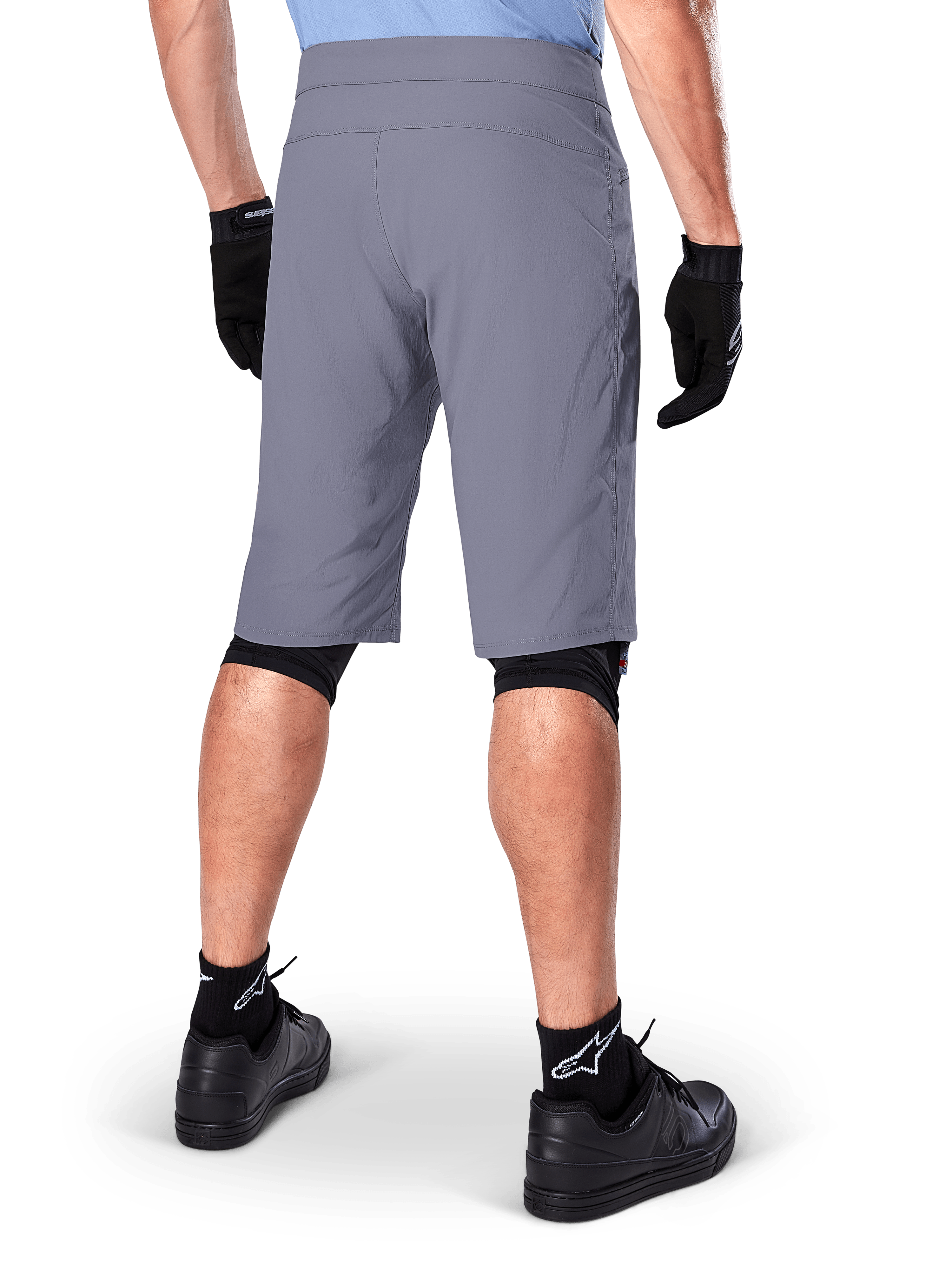 Alpinestars A-Dura Shorts, pantaloncini da mountain bike grigio scuro, vista posteriore su un ciclista, indossati con guanti neri, protezioni ginocchia nere e calze nere con logo Alpinestars, costruzione resistente per il ciclismo fuoristrada