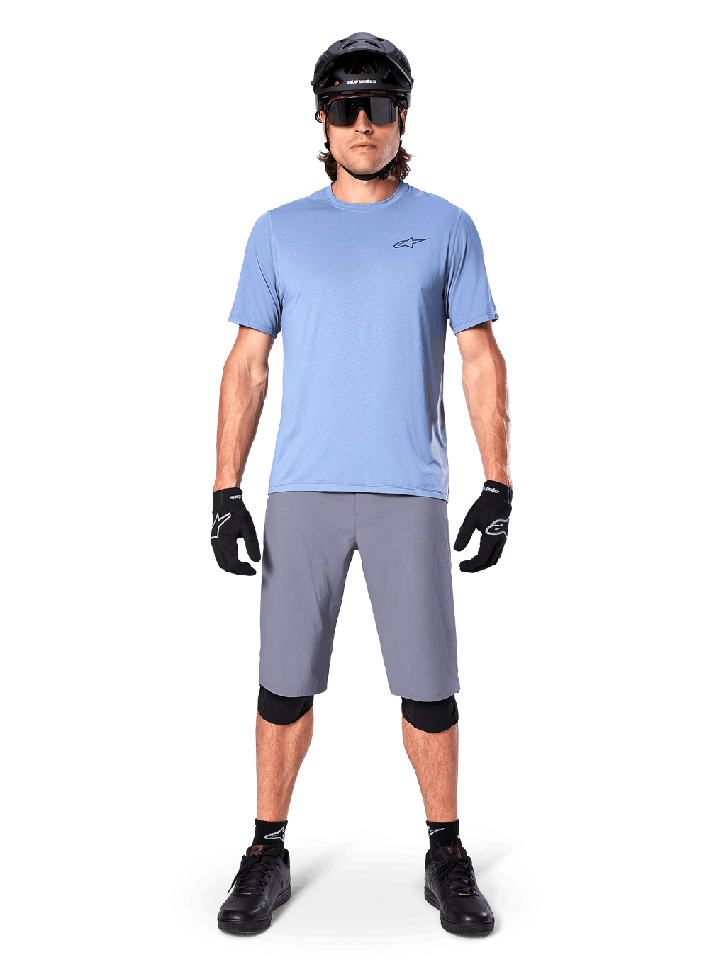 Ciclista uomo che indossa pantaloncini Alpinestars A-Dura in grigio scuro, abbinati a una t-shirt azzurra a maniche corte, casco nero, occhiali da sole e ginocchiere nere, abbigliamento completo da mountain bike per prestazioni off-road.