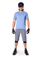 Ciclista uomo che indossa pantaloncini Alpinestars A-Dura in grigio scuro, abbinati a una t-shirt azzurra a maniche corte, casco nero, occhiali da sole e ginocchiere nere, abbigliamento completo da mountain bike per prestazioni off-road.