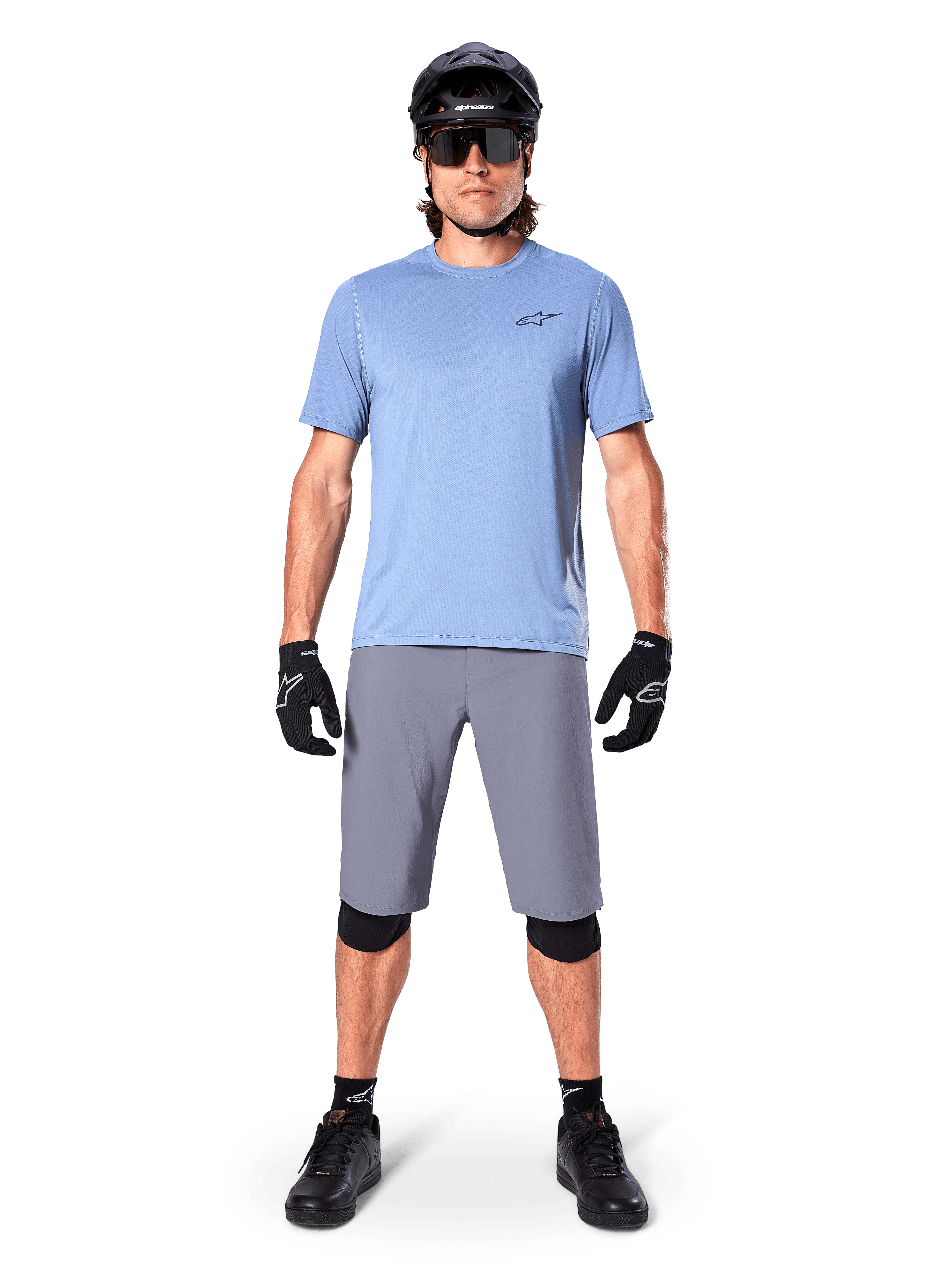 Ciclista uomo che indossa pantaloncini Alpinestars A-Dura in grigio scuro, abbinati a una t-shirt azzurra a maniche corte, casco nero, occhiali da sole e ginocchiere nere, abbigliamento completo da mountain bike per prestazioni off-road.